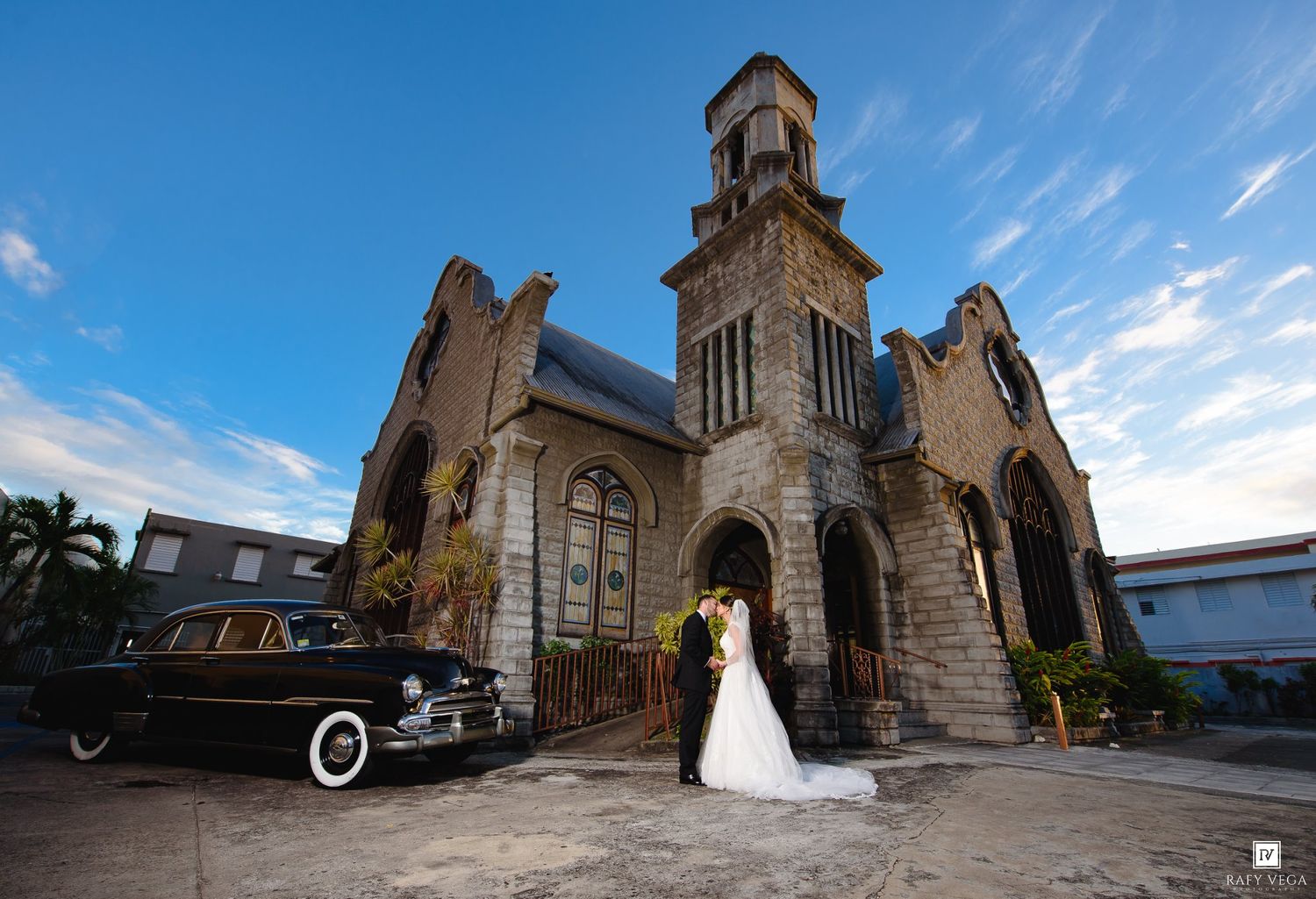 Bride and groom kissing outside a historic stone church with a vintage black car, under a blue sky.
