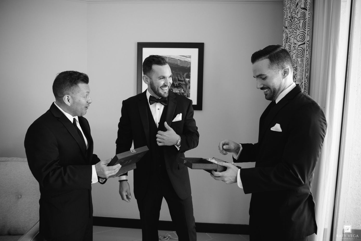 Three groomsmen in black tuxedos laughing and exchanging gifts in black and white wedding photo.