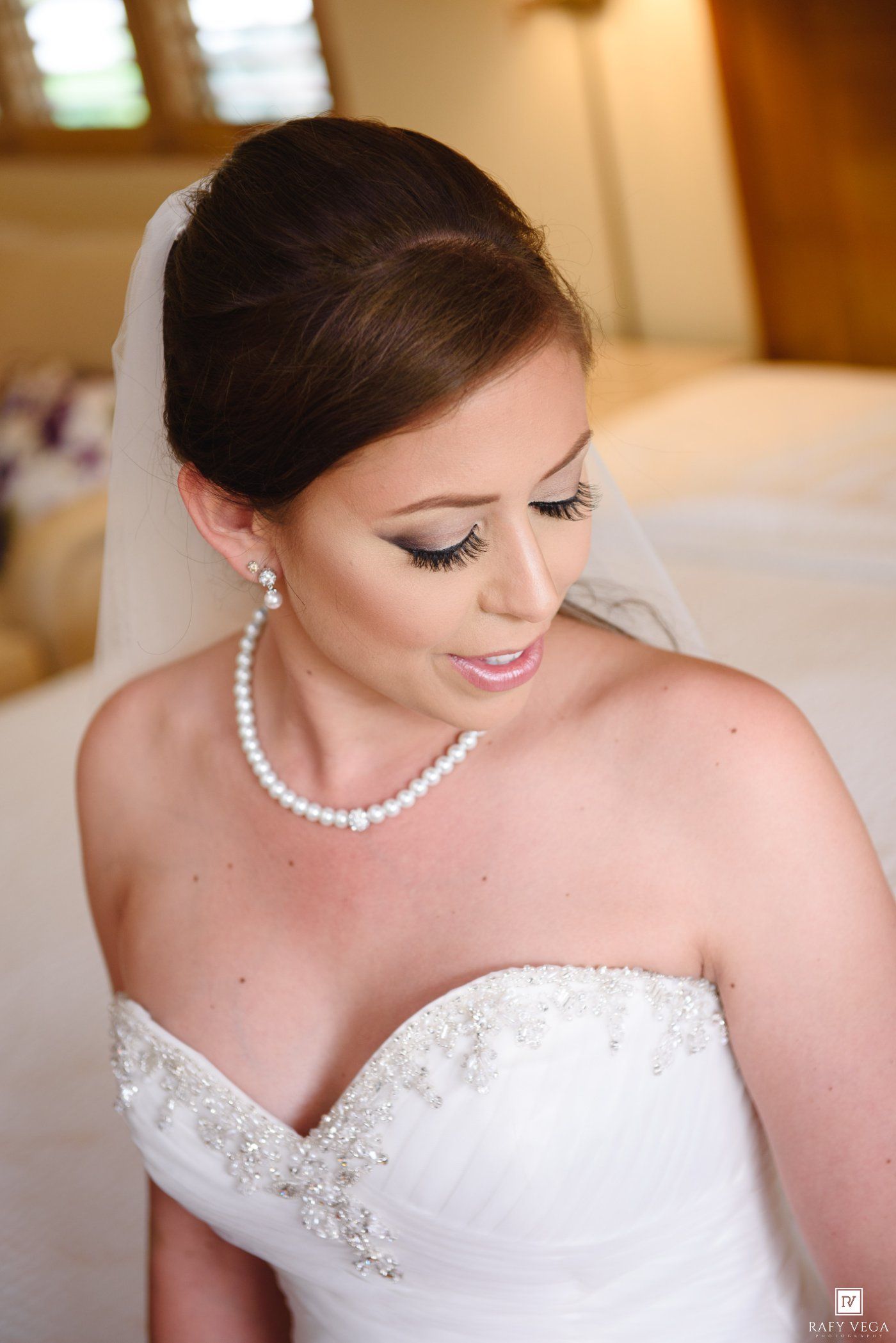 Bride in strapless white gown with pearl necklace and veil, elegant updo hairstyle and natural bridal makeup.
