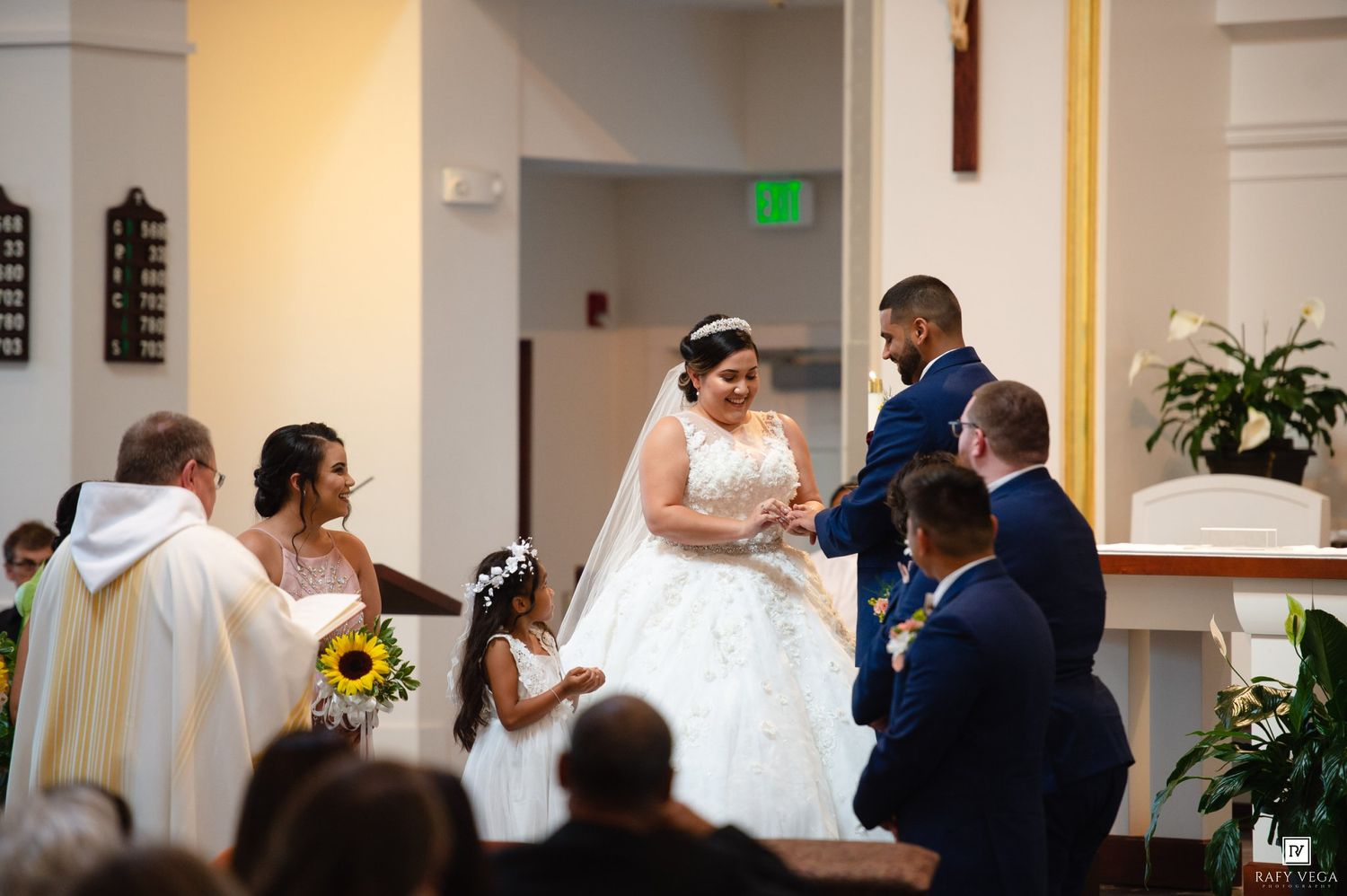 Bride and groom exchange vows at church altar with flower girl and wedding party during Catholic ceremony.