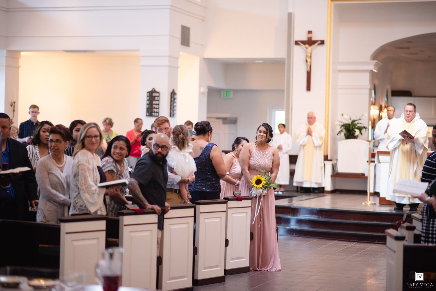 Wedding ceremony in a Catholic church with guests standing in pews, a bridesmaid holding sunflowers, and priest at the altar.