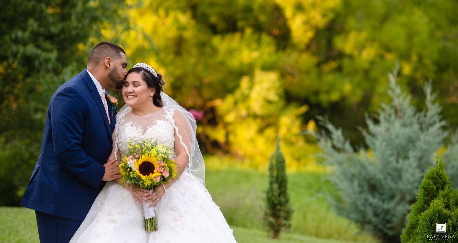 Bride in white gown holds sunflower bouquet as groom in navy suit kisses her cheek in lush green garden.