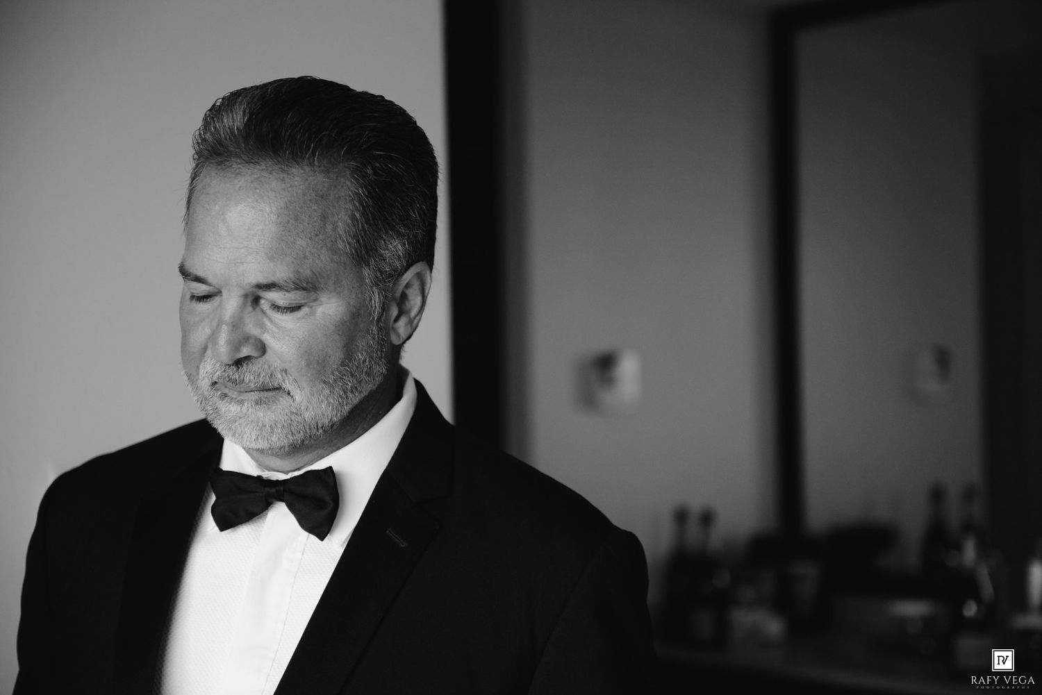 Elegant man in tuxedo with bow tie looking down in moody black and white portrait.