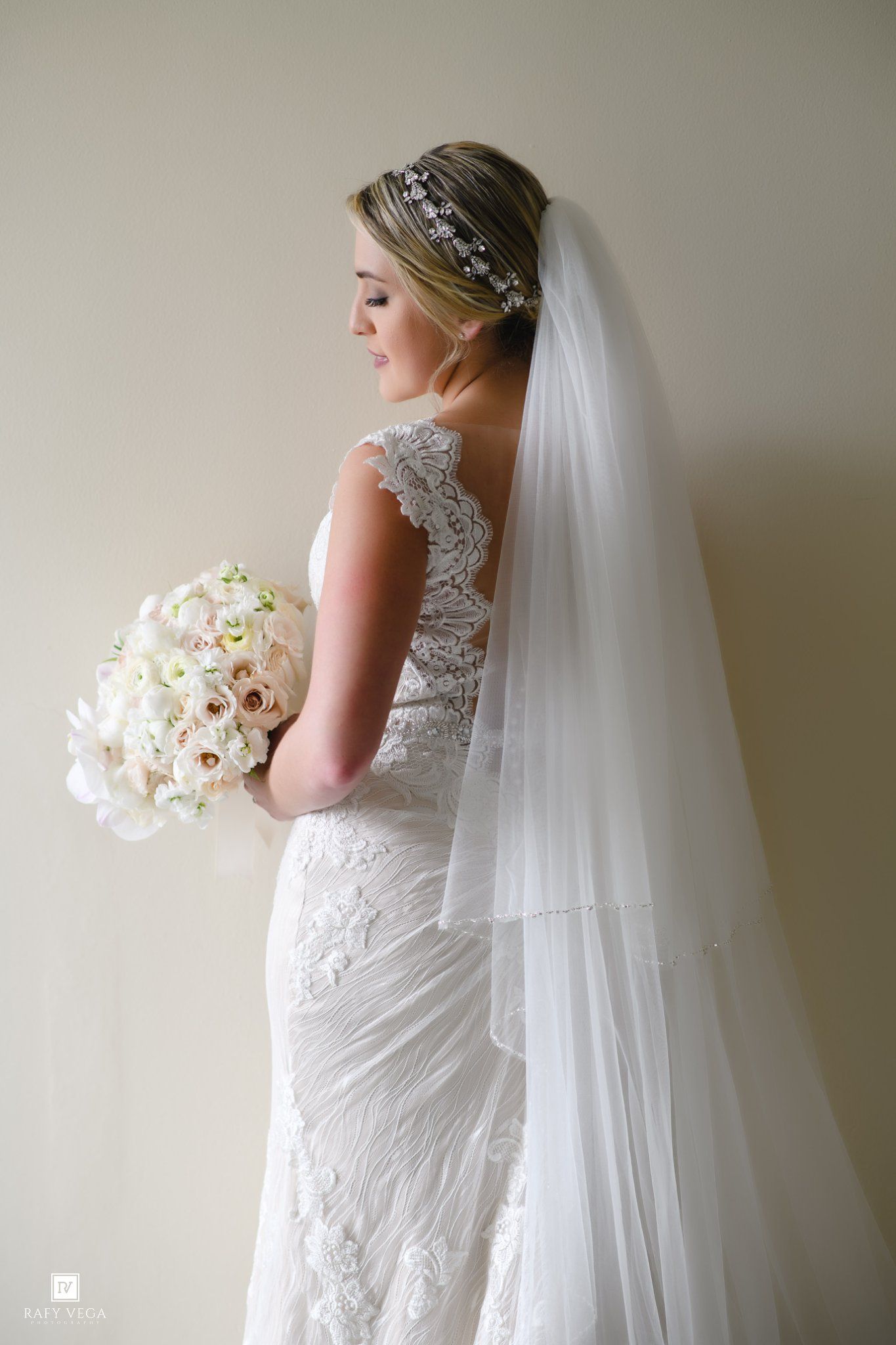 Bride in lace wedding gown with veil and floral headpiece holds white bouquet, elegant bridal portrait.