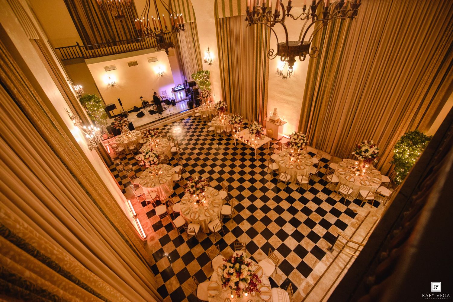 Elegant ballroom with black and white checkered floor, candlelit tables, and warm golden drapery for a formal event.