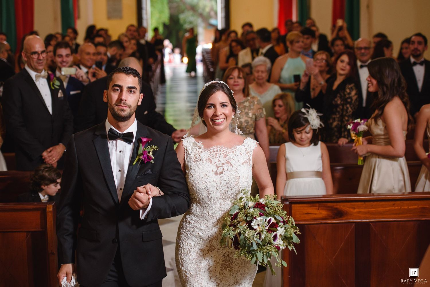 Bride and groom walking down the aisle in a church, bride holding a lush bouquet, guests seated in wooden pews.