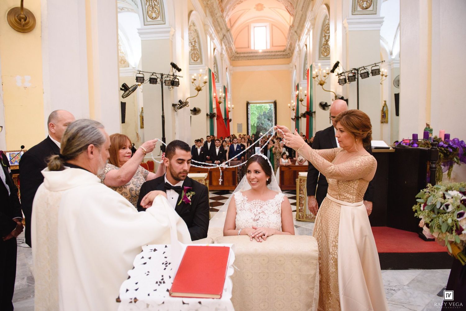 Priest performs traditional wedding ceremony blessing over bride and groom seated at altar in church.
