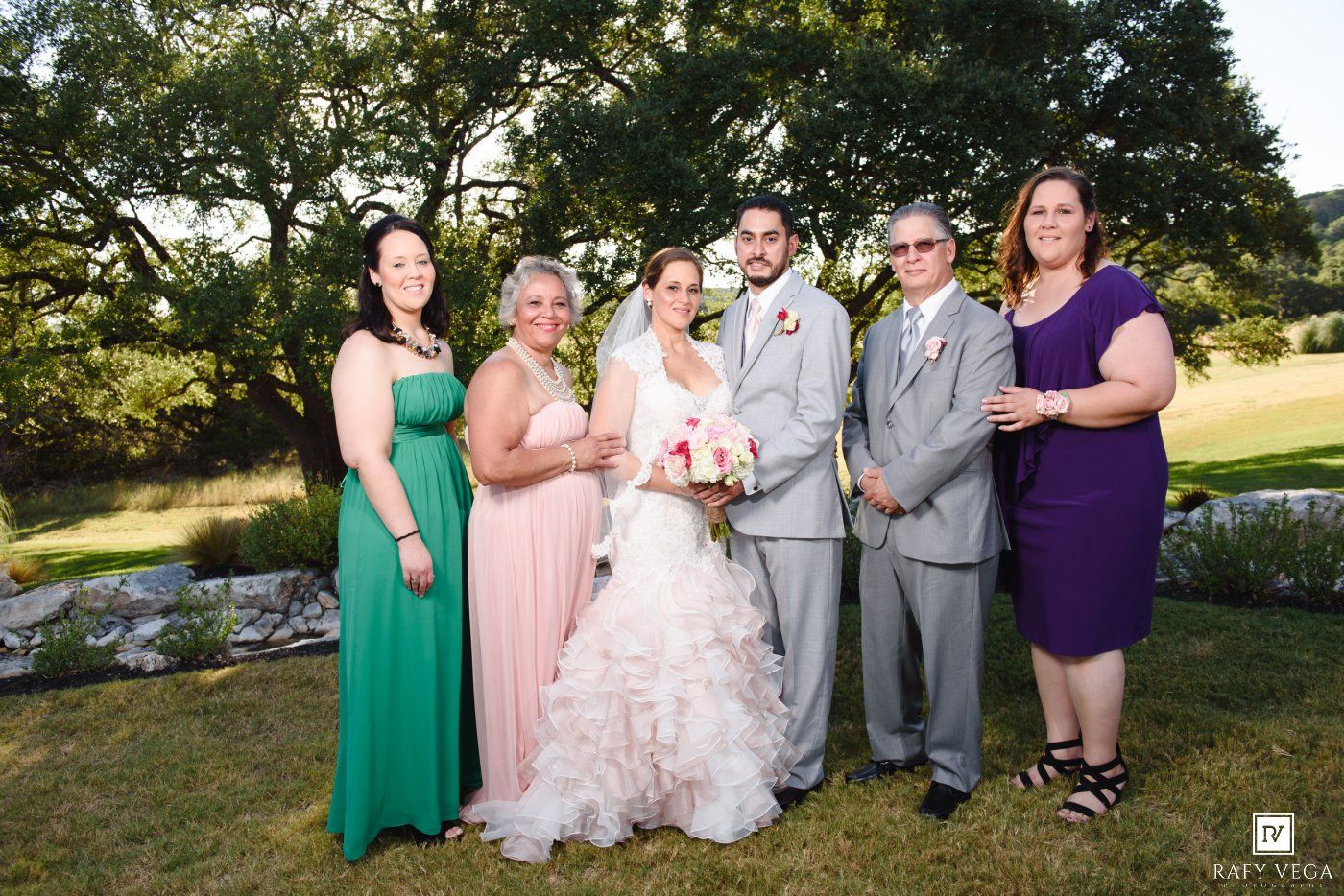 Wedding party posing outdoors, bride in white gown with bridesmaids in green and purple dresses, groom in gray suit.