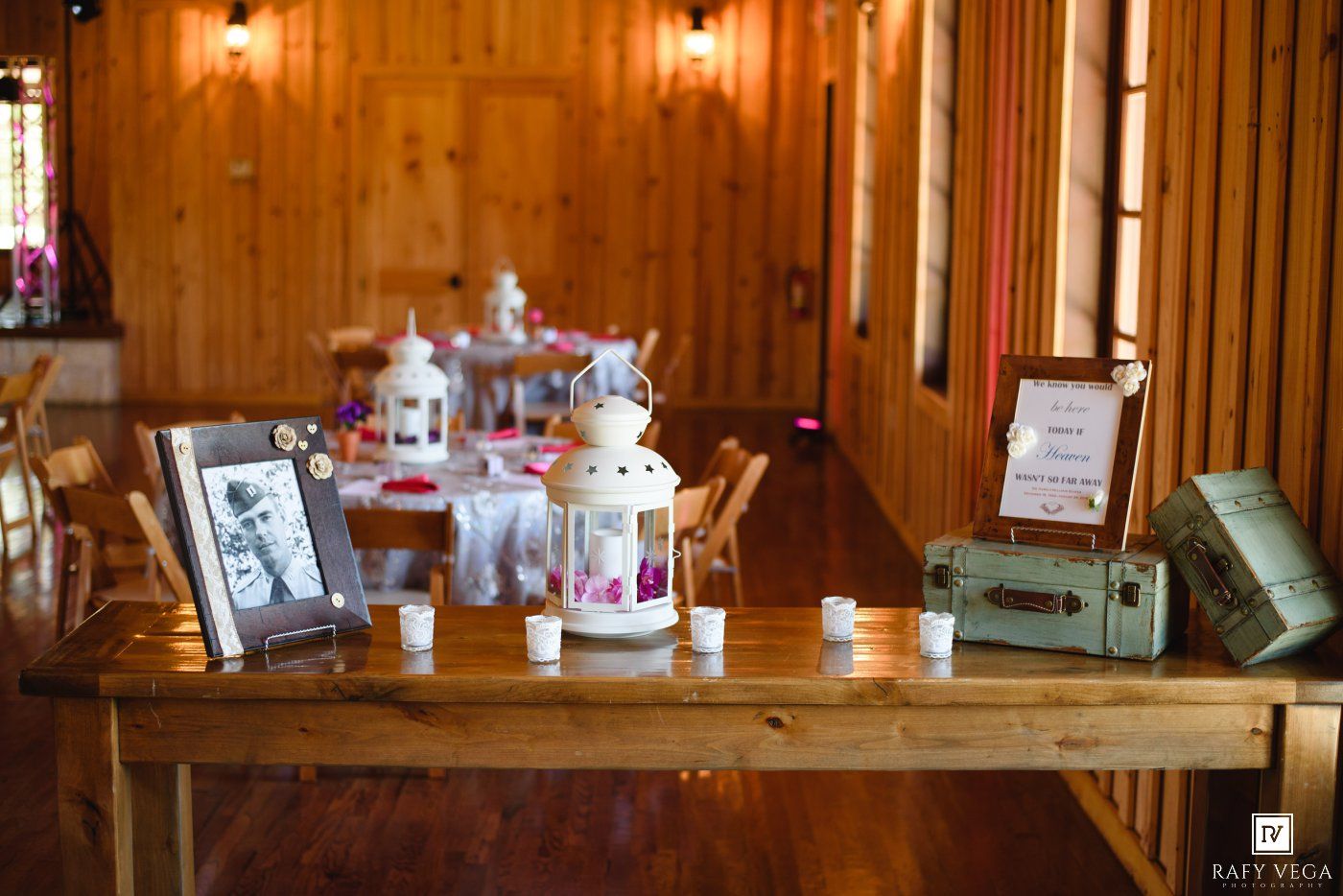 Rustic wooden wedding welcome table with lantern centerpiece, framed photos, candles, and vintage suitcases in warm venue.