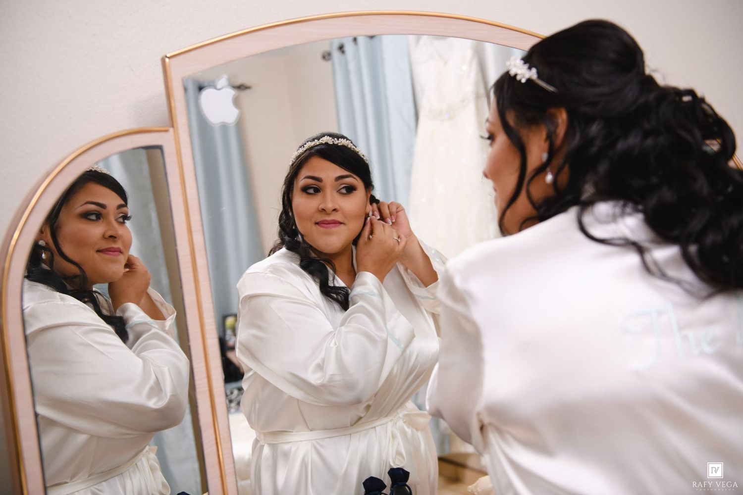 Bride in white robe adjusting earrings before wedding, reflected in elegant tri-fold mirror.