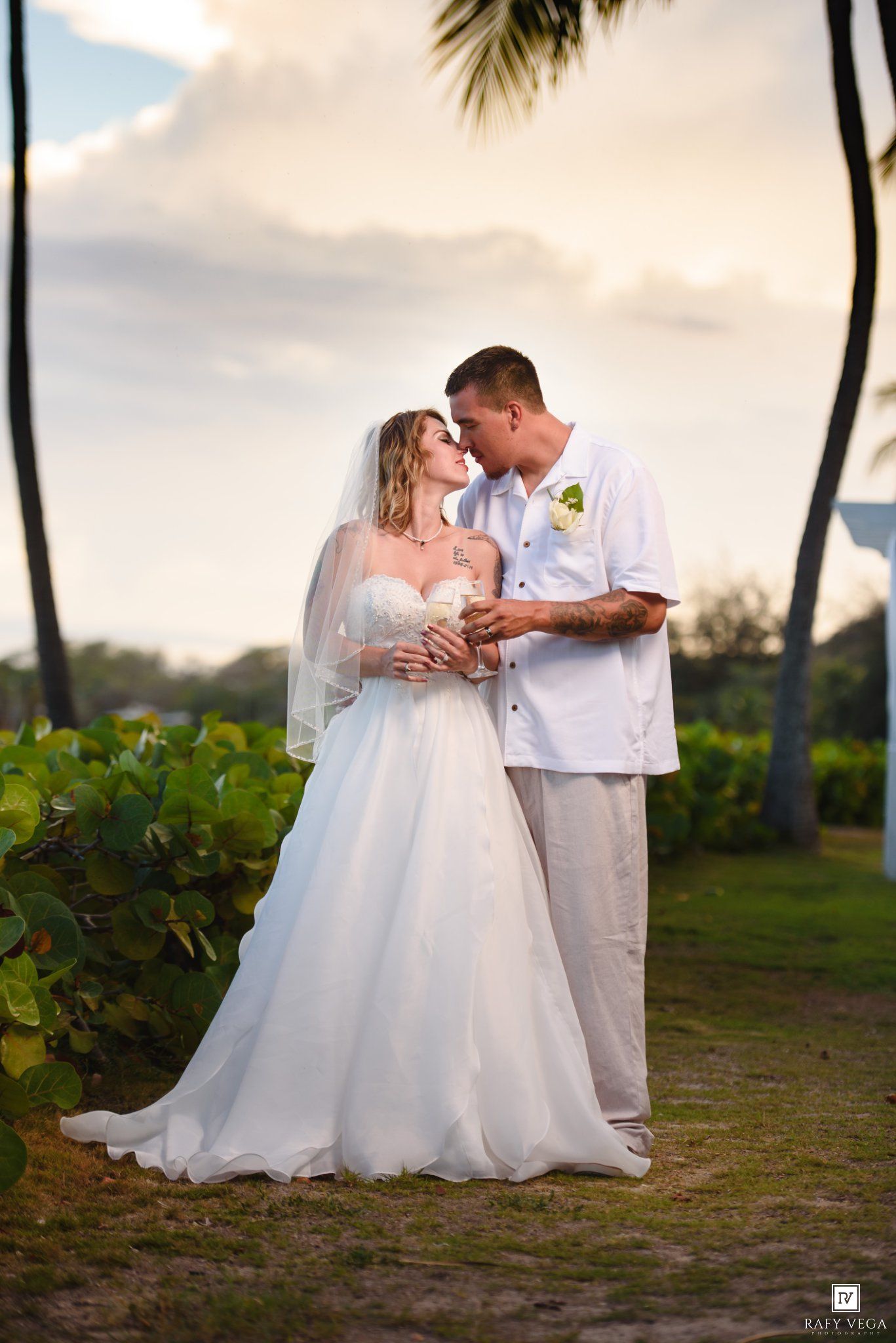 Bride and groom share a romantic moment at a tropical beach wedding, surrounded by palm trees at sunset.