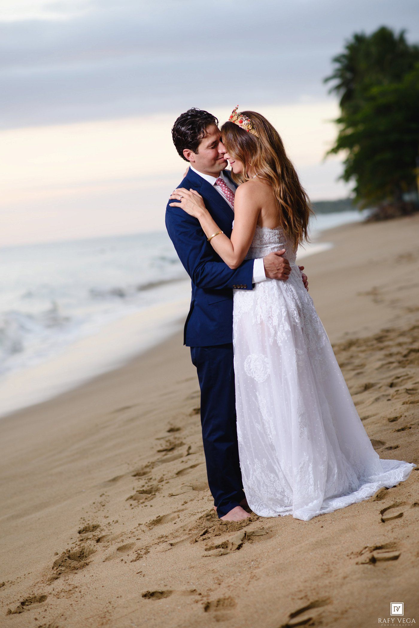 Bride and groom embrace romantically on a sandy beach at sunset, she in a white gown, he in a navy suit.