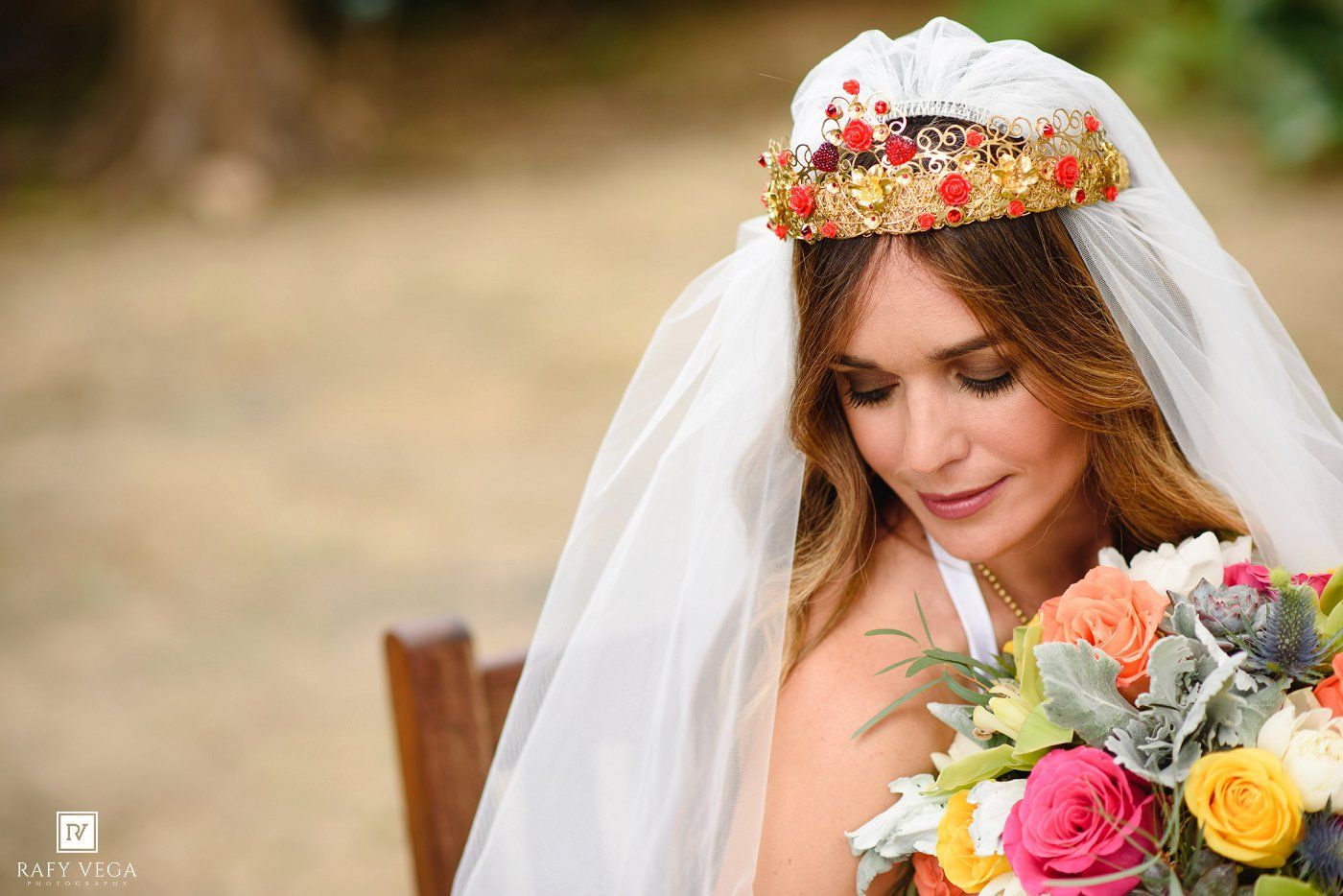 Bride wearing gold floral crown and white veil holds colorful bouquet of roses outdoors on her wedding day.