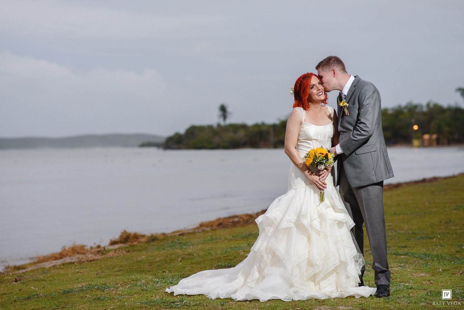 Bride in white gown with red hair holds bouquet beside groom in gray suit near waterfront on wedding day.
