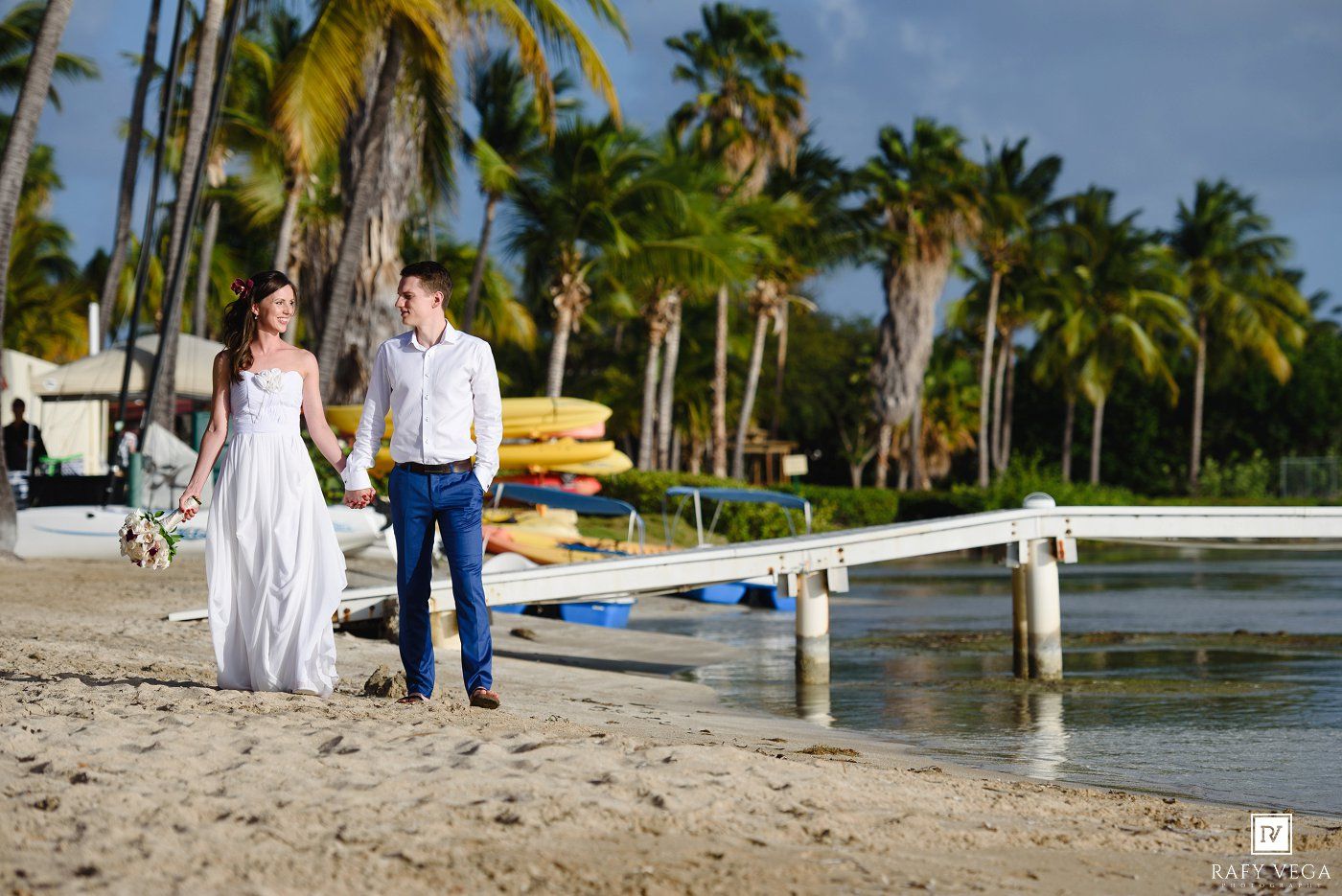 Couple walks hand-in-hand on tropical beach near wooden dock, palm trees and colorful kayaks in background.