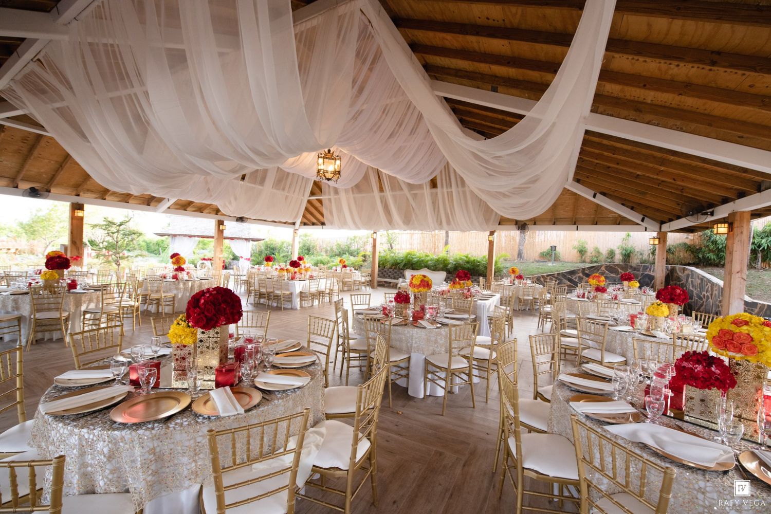 Elegant outdoor wedding reception venue with white draped ceiling, gold chiavari chairs, and vibrant floral centerpieces.