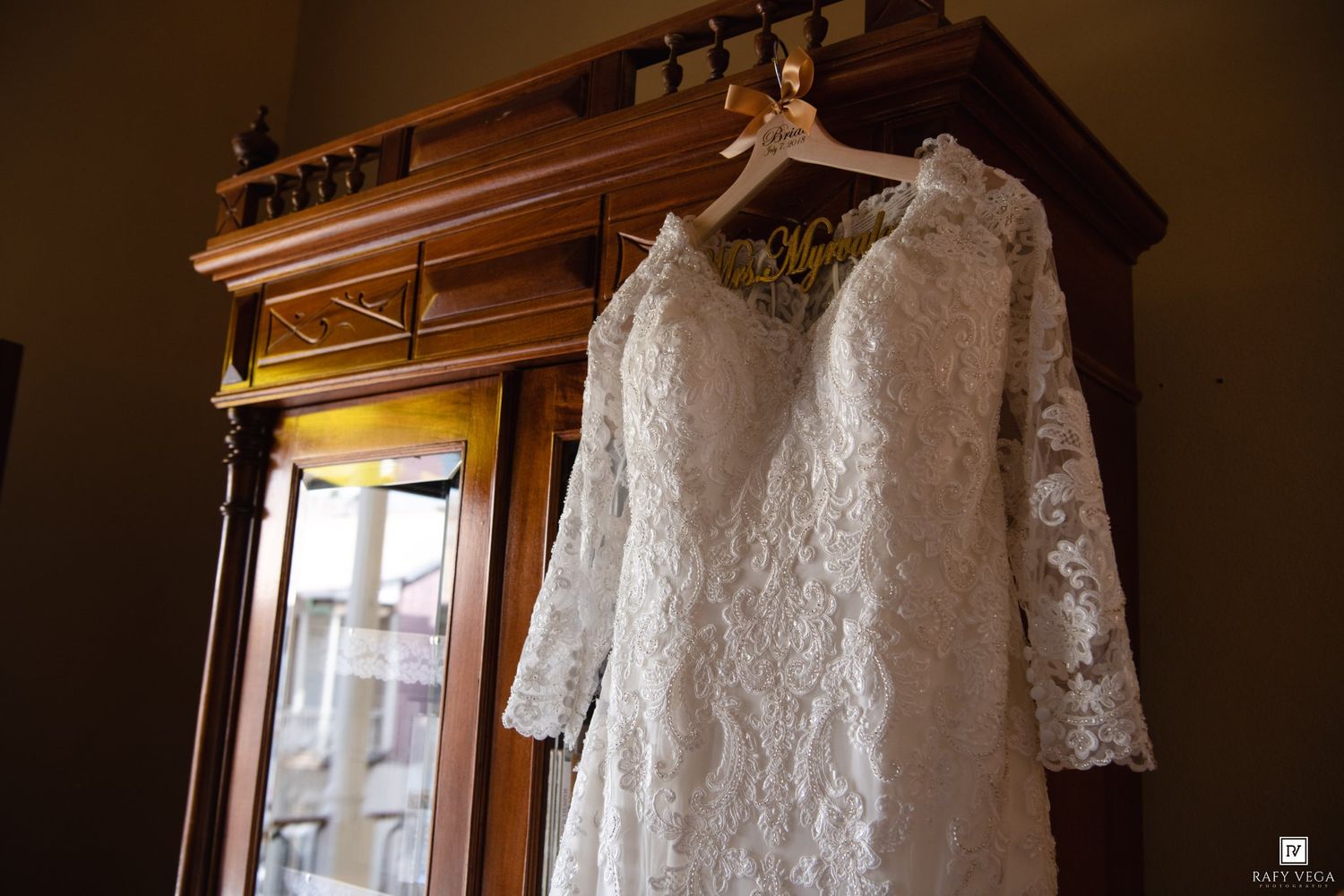 Elegant lace wedding dress with long sleeves hanging on a wooden armoire hanger in a warmly lit room.