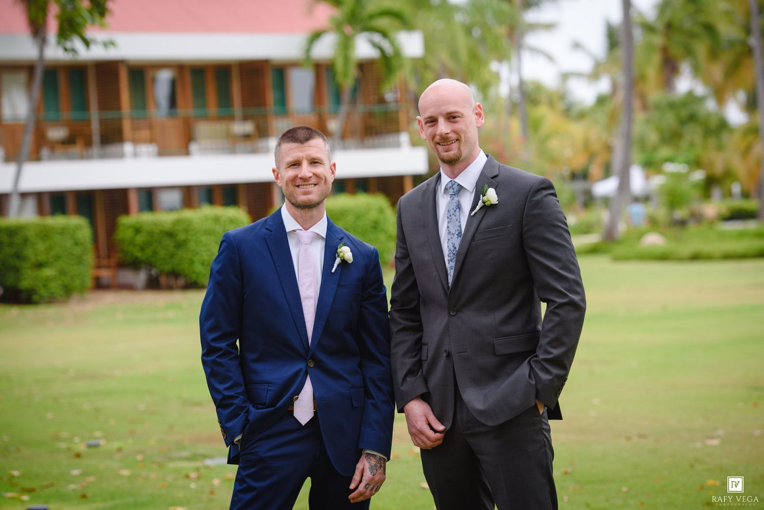 Two grooms in suits smiling outdoors at a tropical wedding venue with lush greenery and a building in the background.