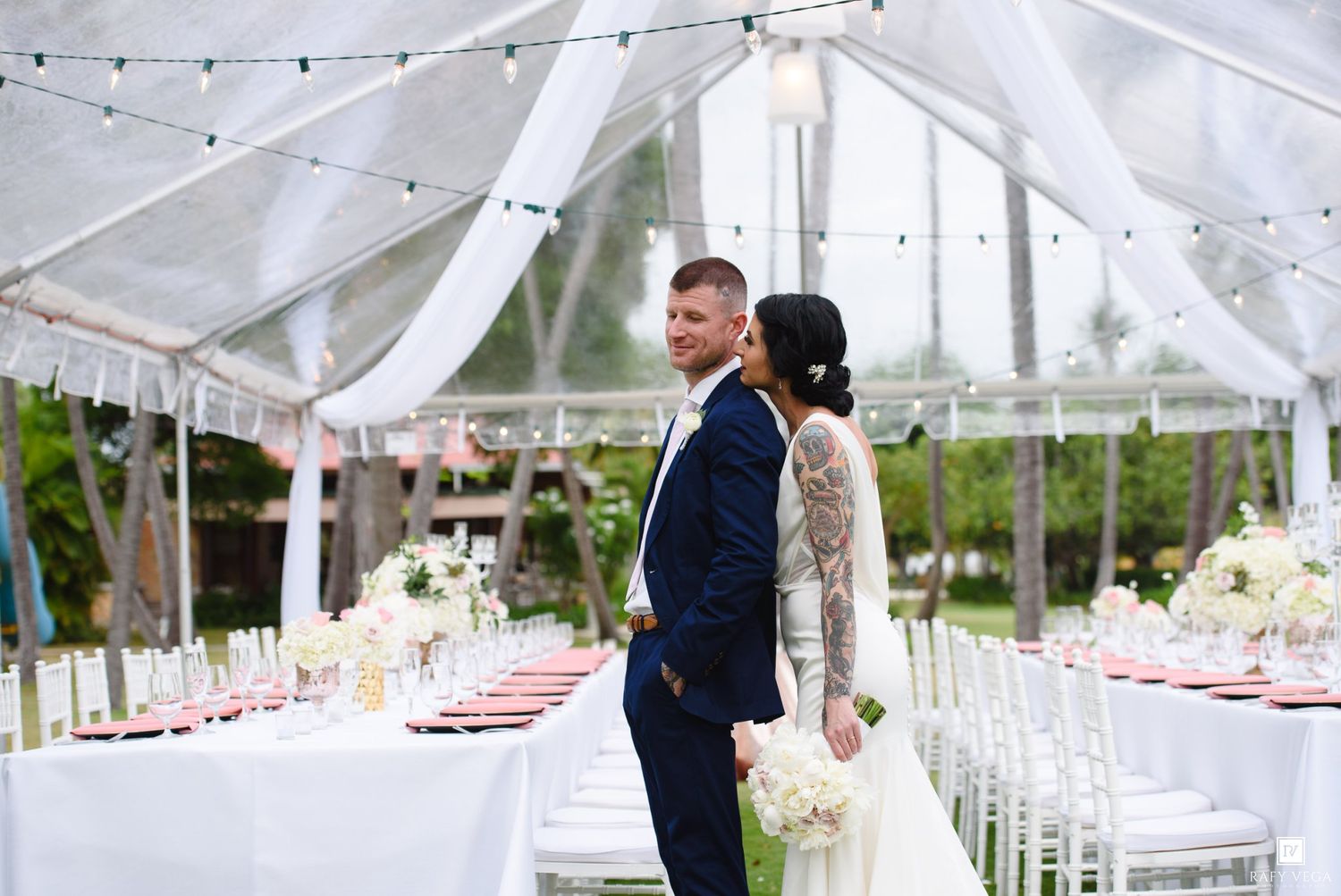 Bride and groom pose under elegant white tent with floral centerpieces at outdoor wedding reception.