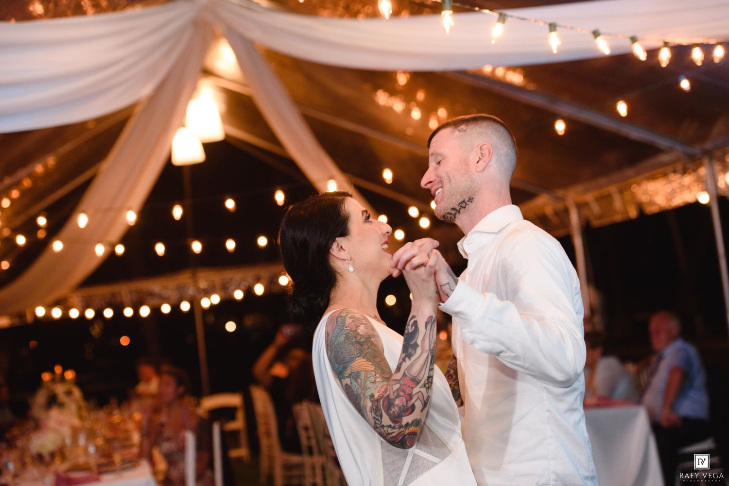 Bride and groom share first dance under string lights in elegant draped tent at outdoor wedding reception.