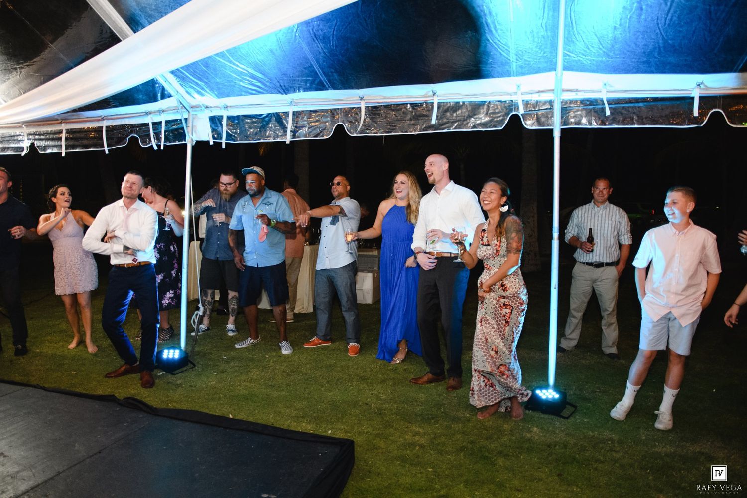 Guests socializing and dancing under a white tent at an outdoor evening party with blue ambient lighting.