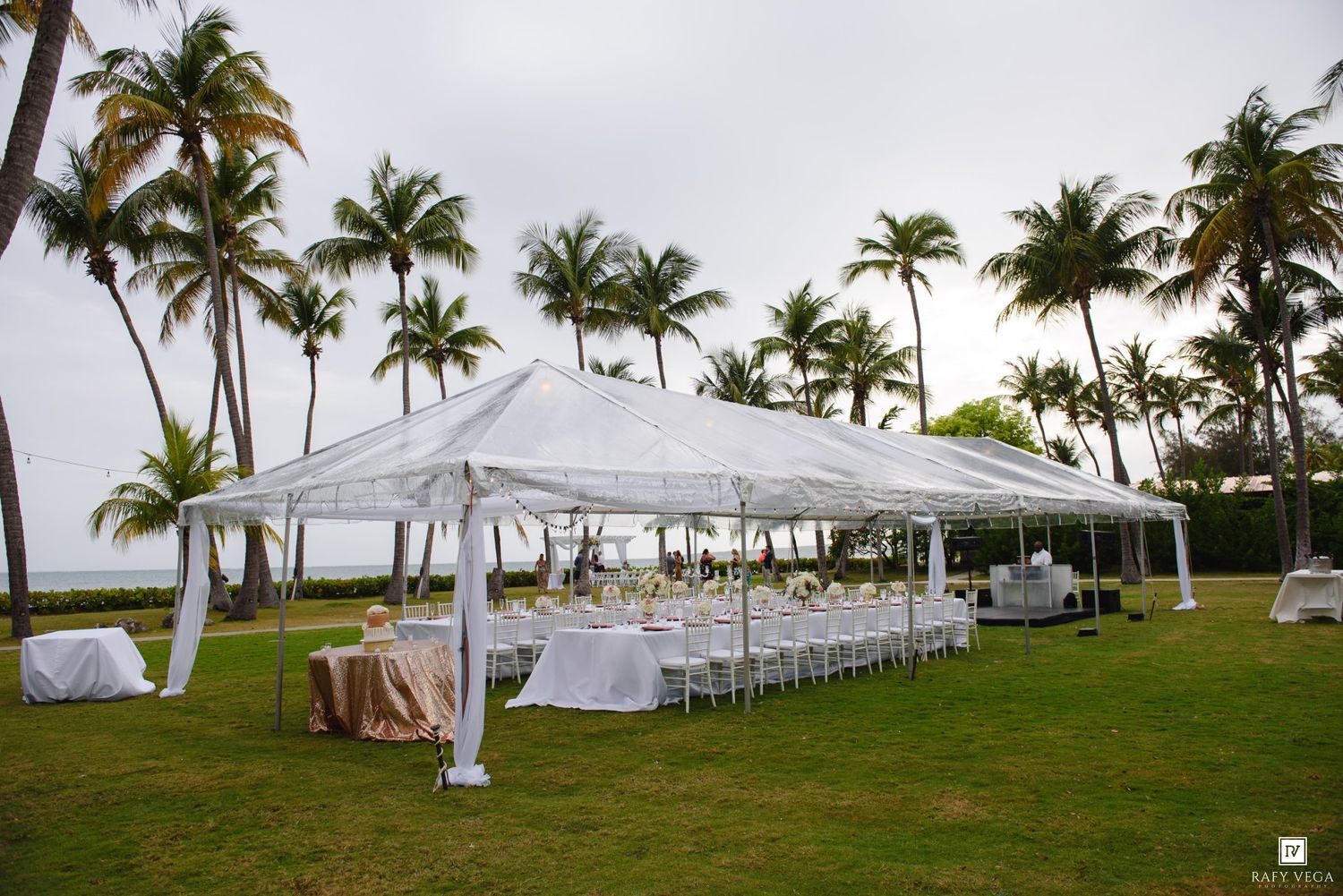 Elegant outdoor wedding reception tent setup on tropical lawn with palm trees and ocean views.