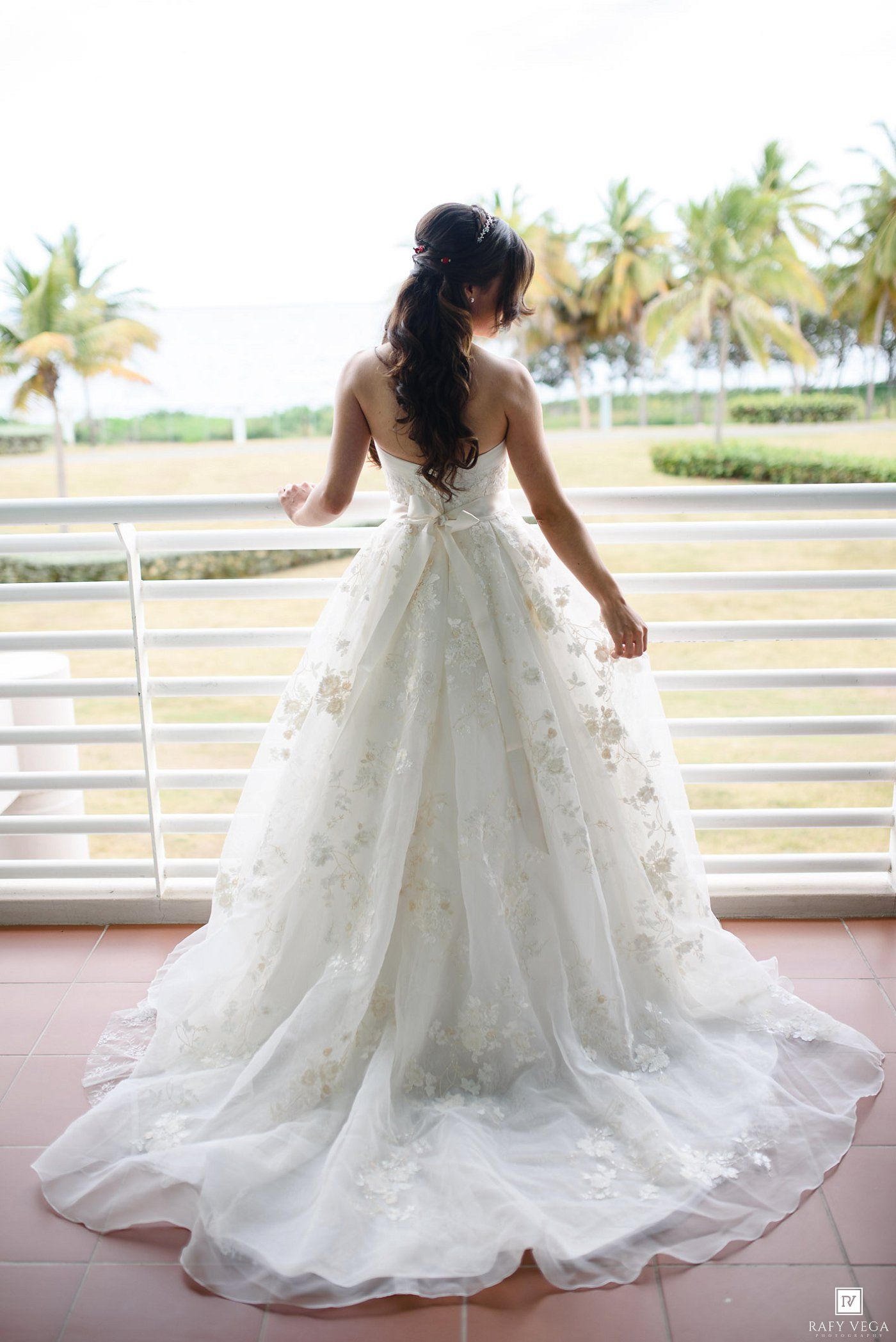 Bride in a stunning white floral ball gown wedding dress stands on a balcony overlooking tropical palm trees.