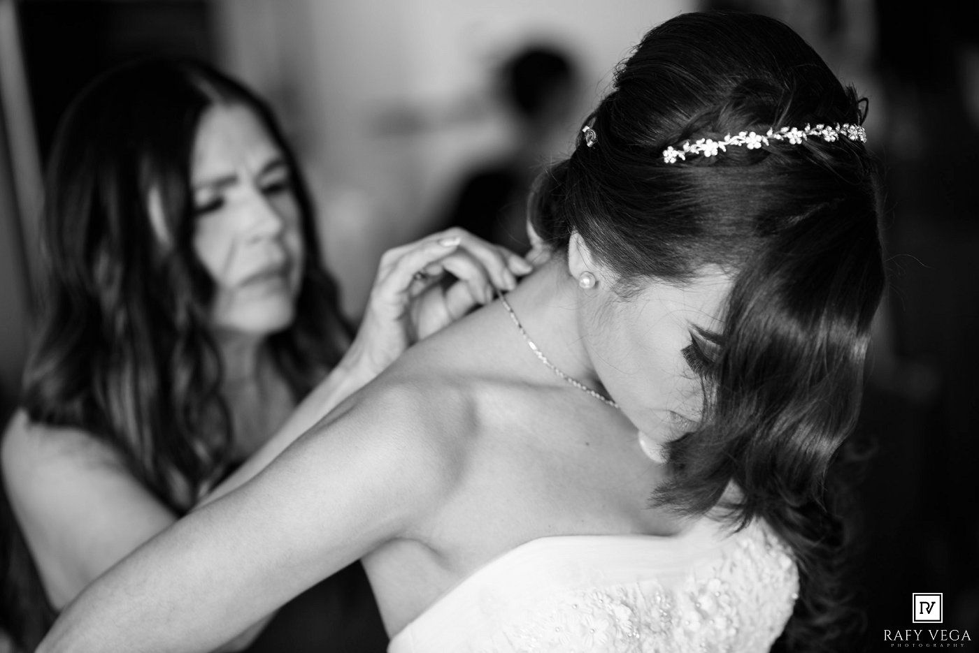 Black and white photo of a bride having her necklace fastened by a bridesmaid on her wedding day.