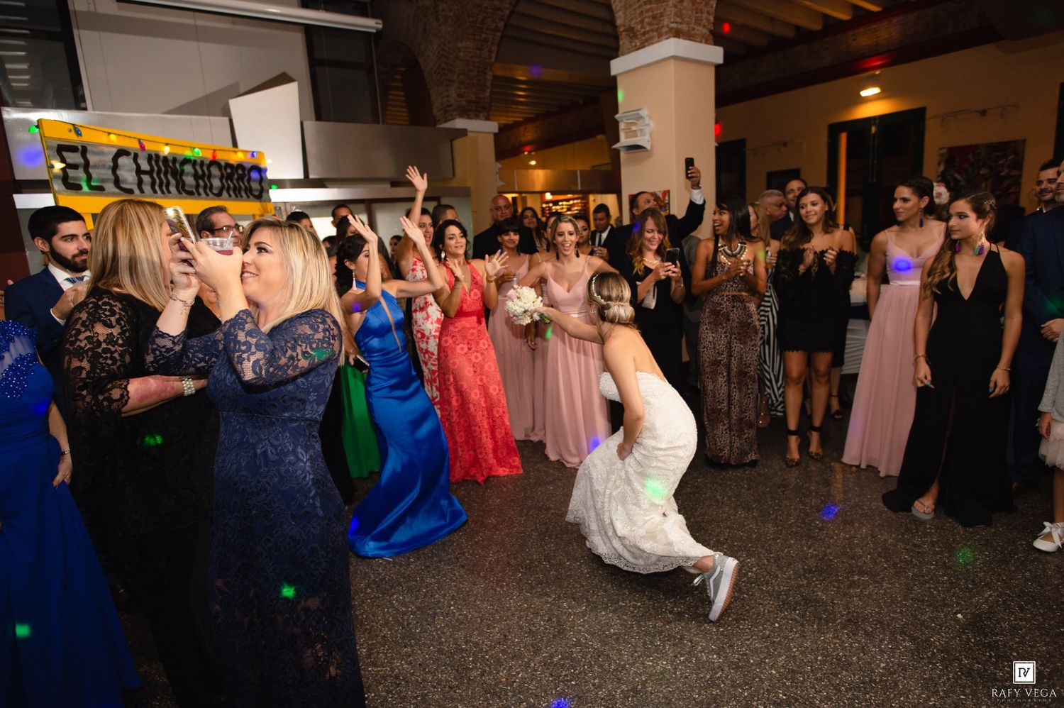 Bride tossing bouquet to excited bridesmaids in colorful gowns at a lively indoor wedding reception.