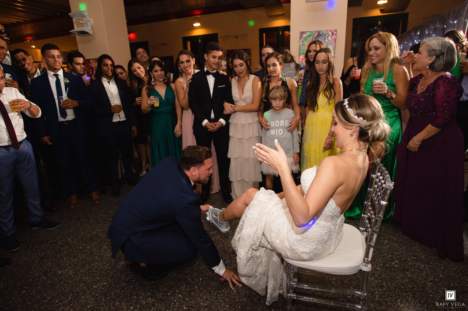 Groom kneeling to place garter on bride seated in chair at wedding reception with guests watching.