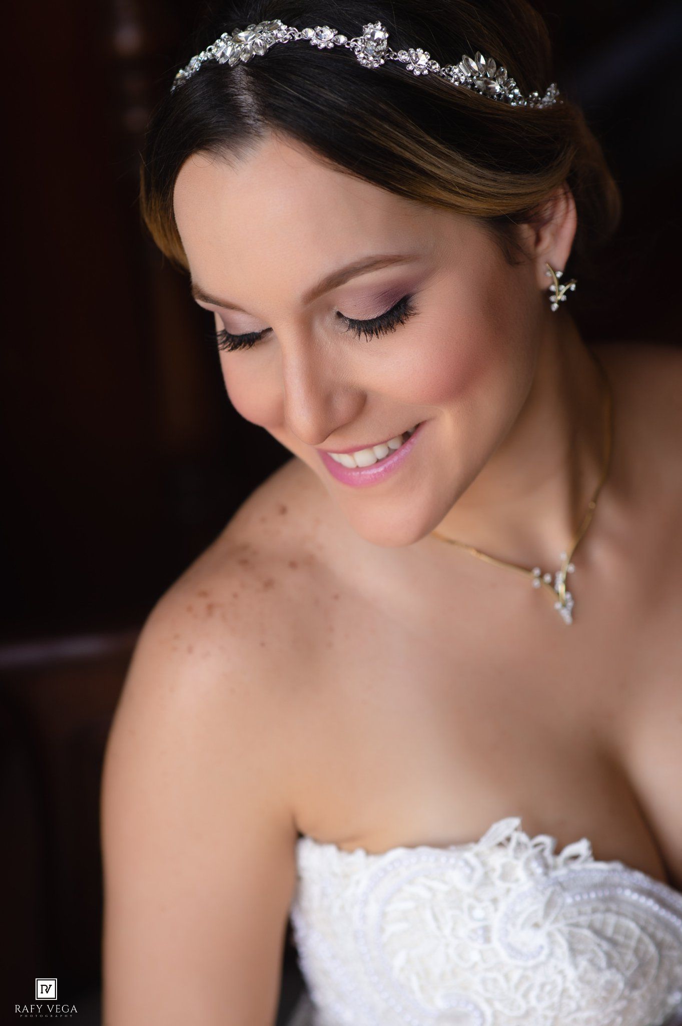 Smiling bride wearing a crystal tiara, pearl earrings, and strapless lace wedding gown with elegant bridal makeup.