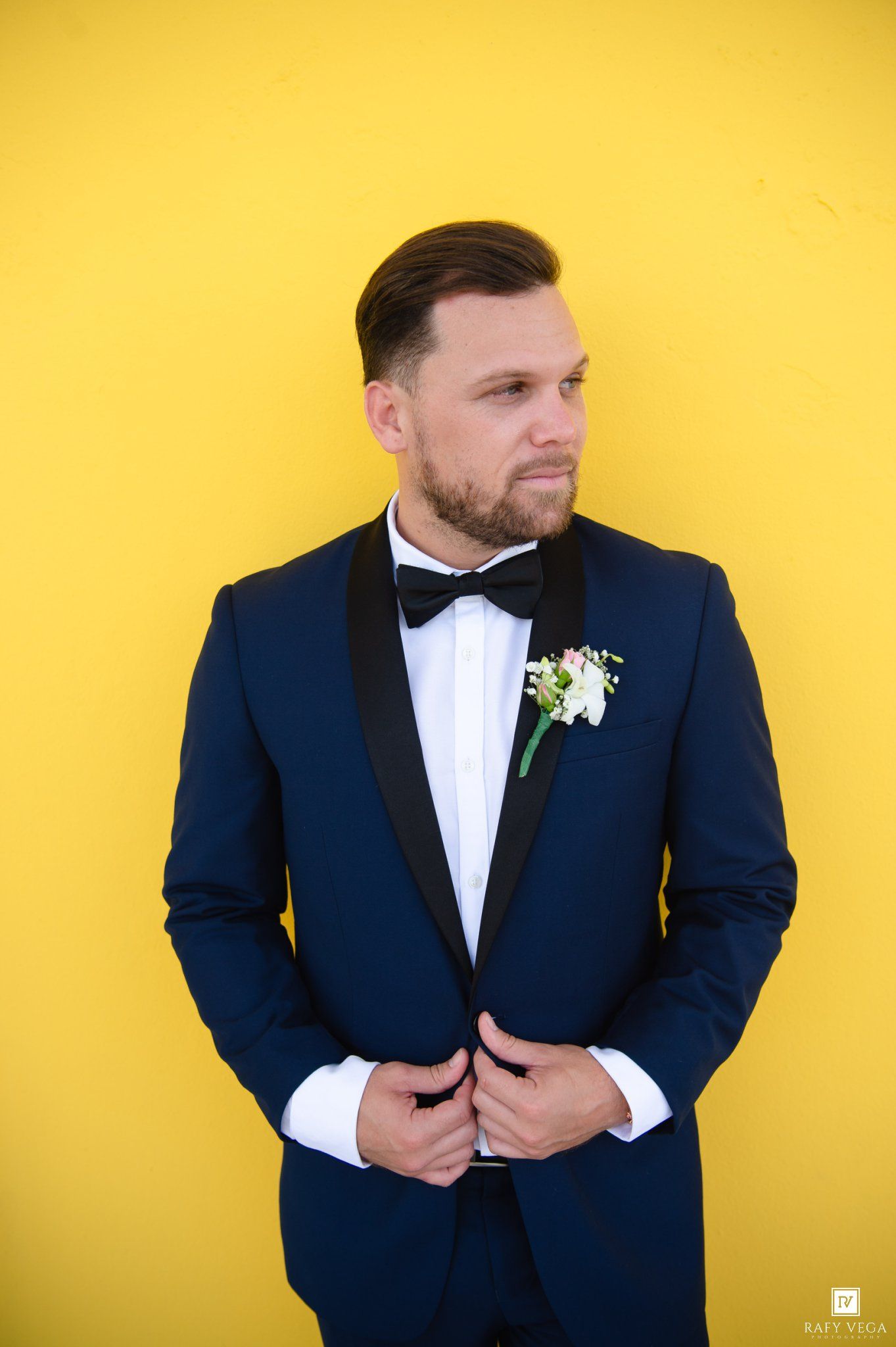 Groom in navy blue tuxedo with bow tie and boutonniere posing against bright yellow wall.