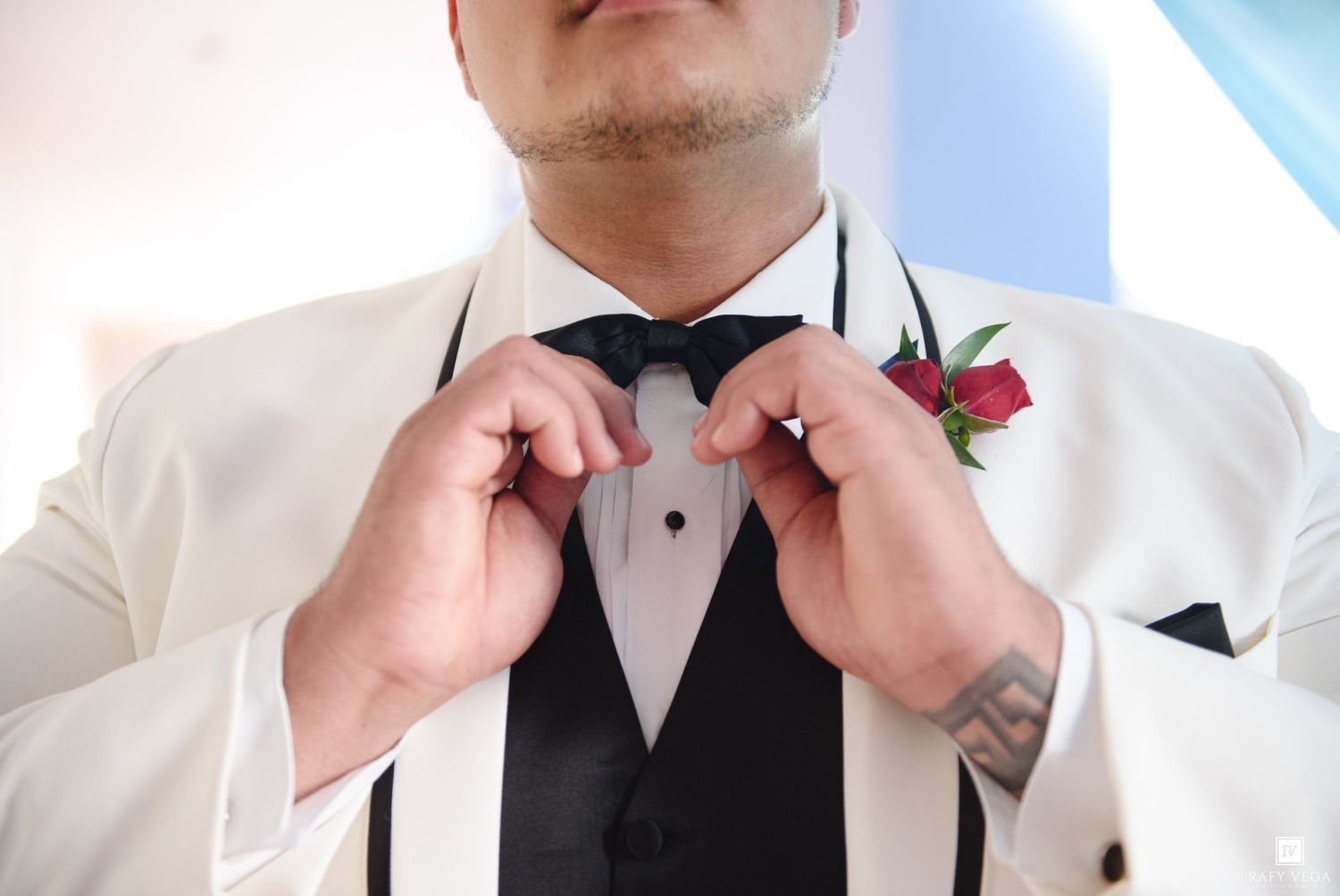 Groom in white tuxedo jacket adjusting black bow tie with floral boutonniere and tattoo visible.