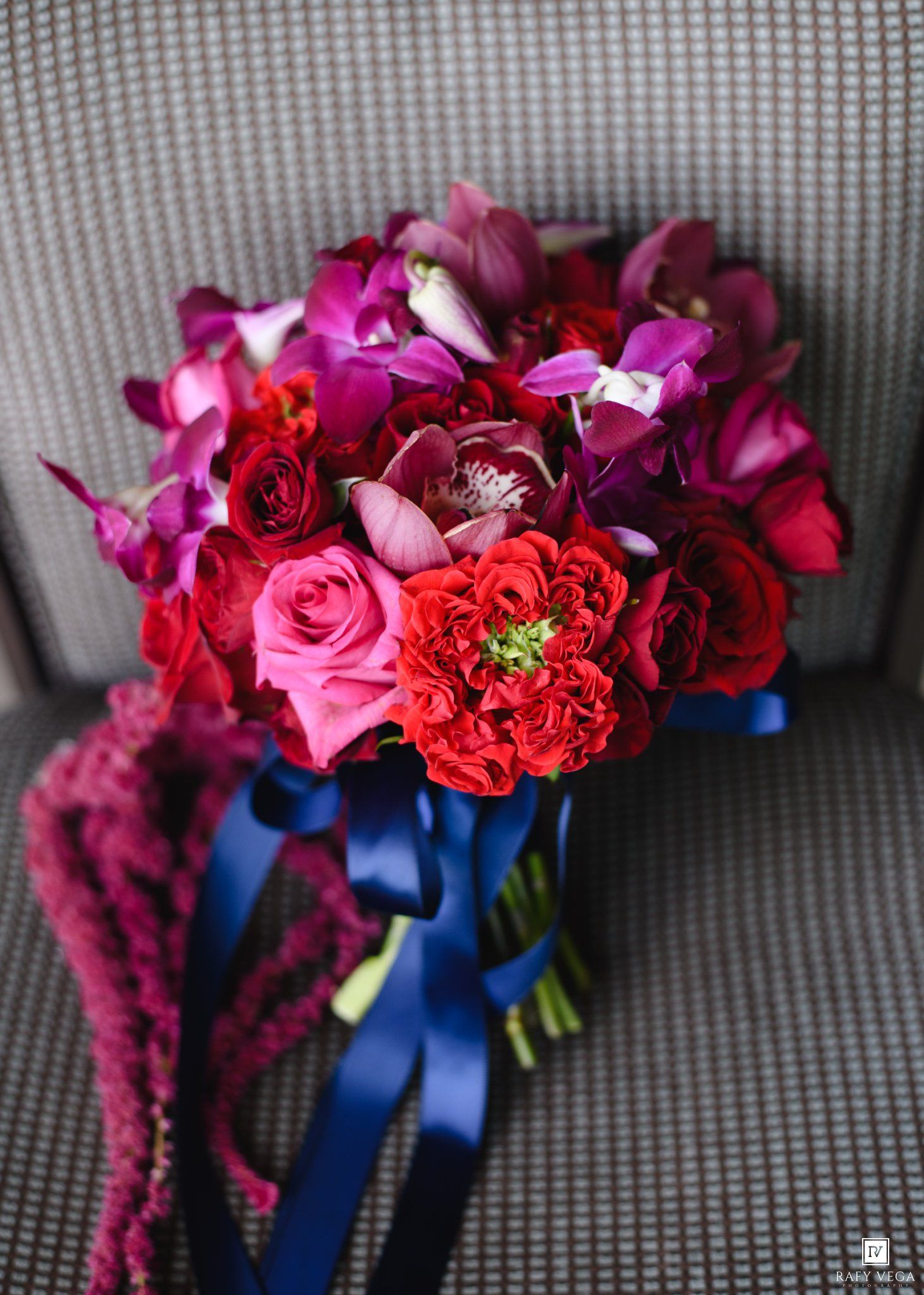 Vibrant bridal bouquet of red roses, pink blooms, and purple orchids tied with a navy blue ribbon.