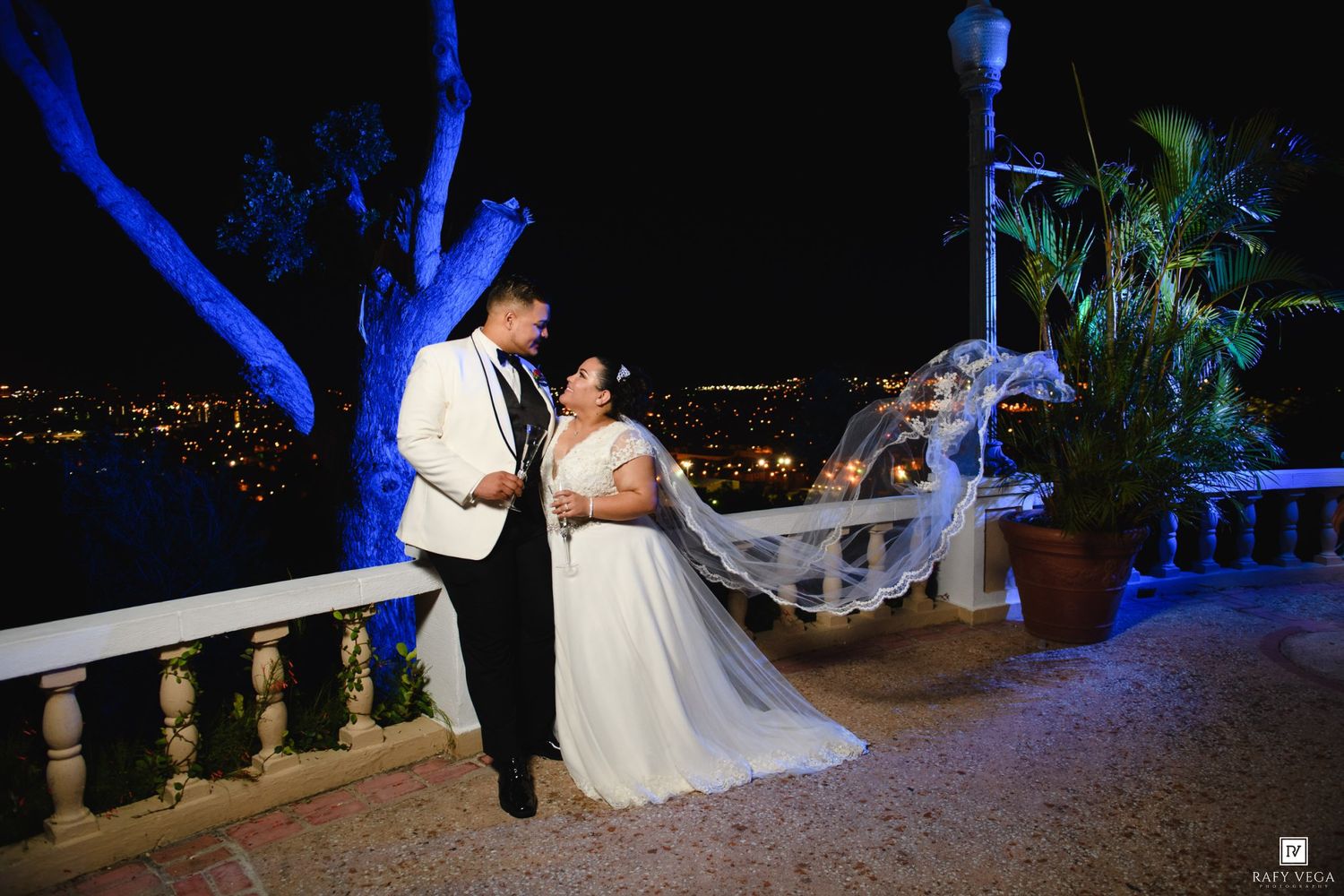 Bride and groom share romantic moment on outdoor terrace at night with city lights in background, veil flowing in breeze.