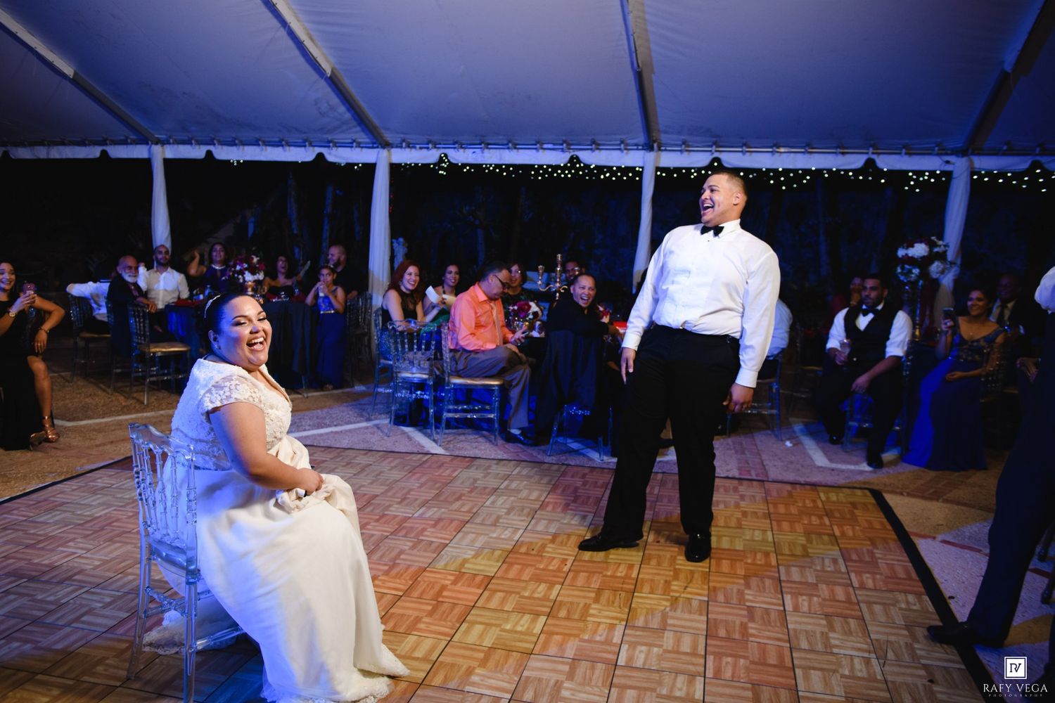 Bride in white gown sits as groom performs during wedding reception dance under tent lights.