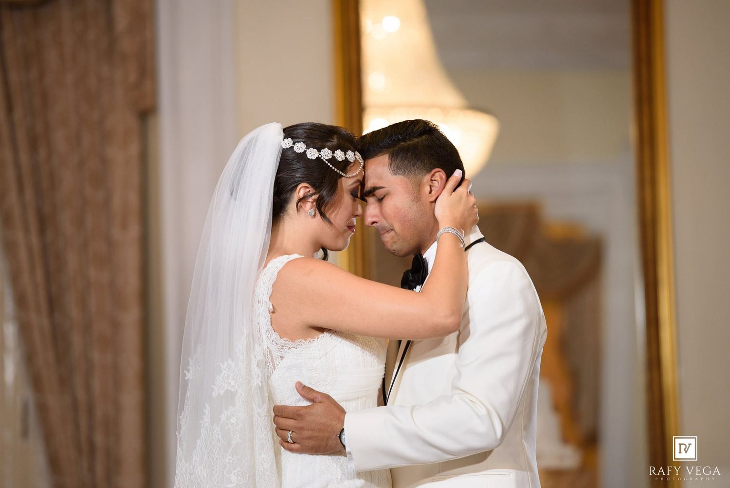 Bride and groom share an intimate forehead touch moment in elegant wedding attire, captured in a warmly lit room.