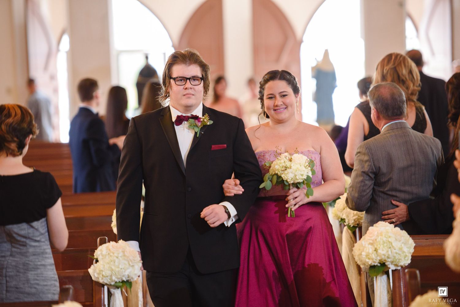 Groomsman in black tux escorts bridesmaid in magenta gown down church aisle at elegant wedding ceremony.