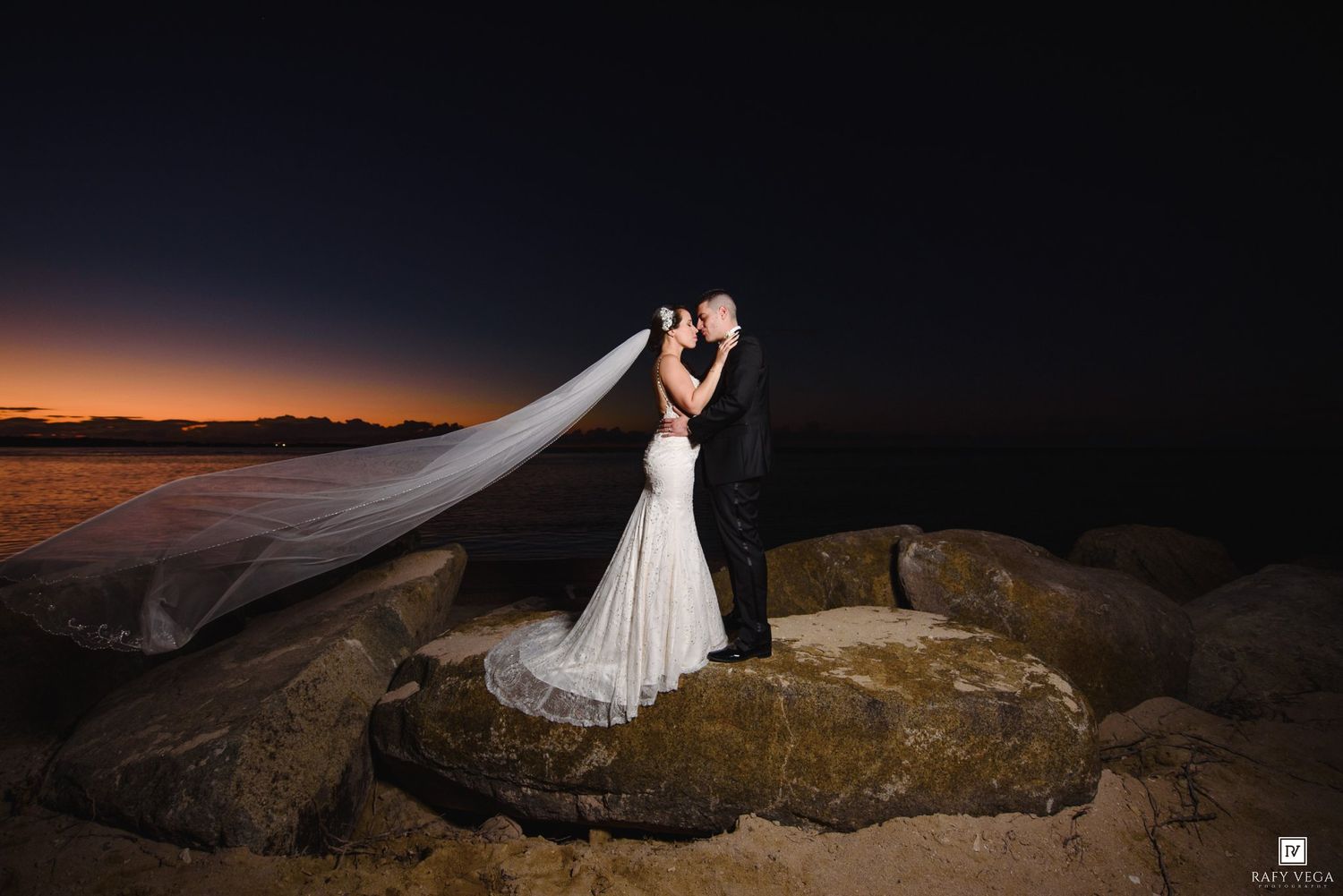 Bride and groom share a romantic kiss on rocky shoreline at sunset, her veil flowing dramatically in the wind.