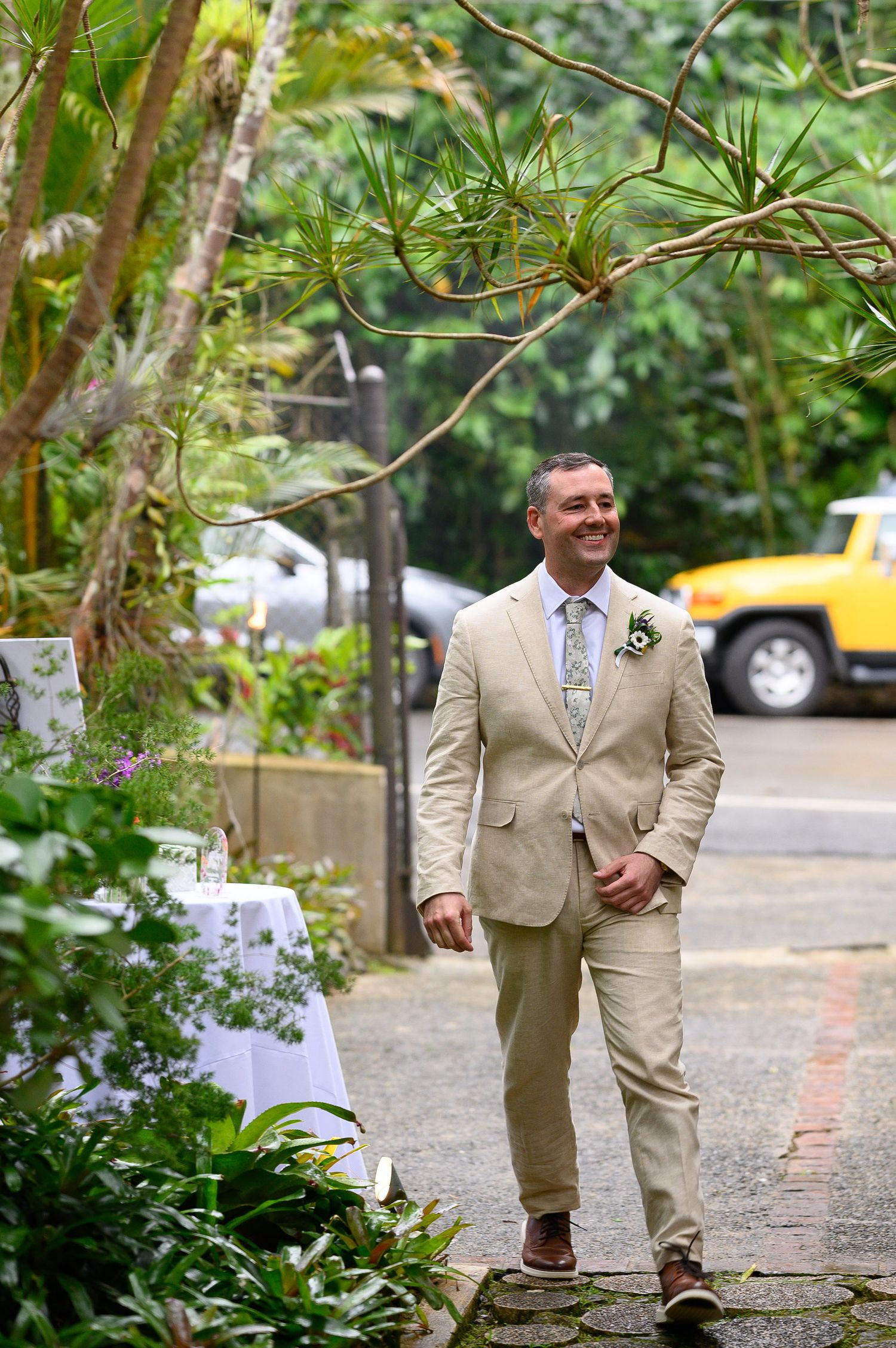 Smiling groom in tan suit with boutonniere walks down lush tropical garden pathway at outdoor wedding ceremony.