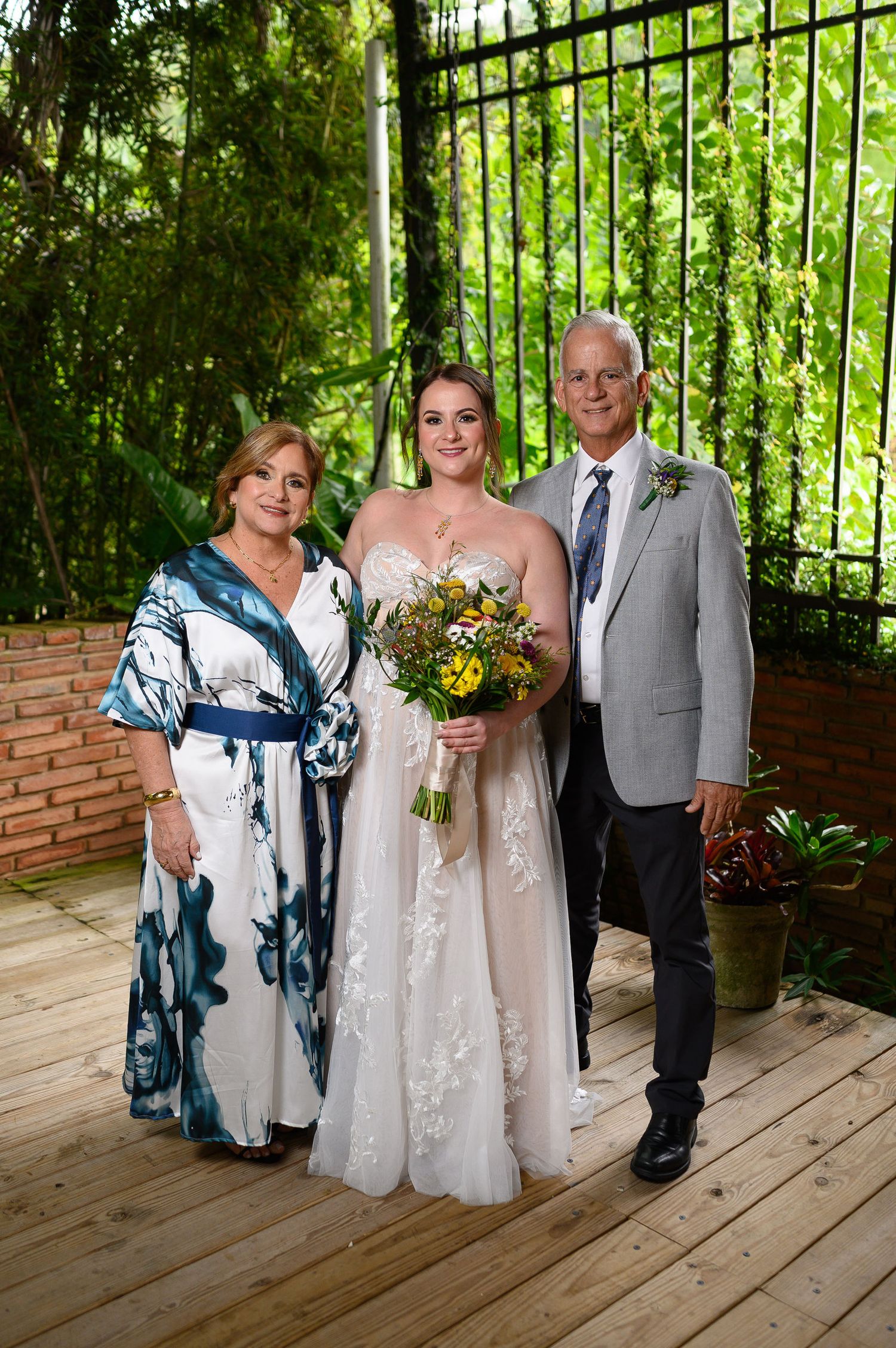 Bride in white gown holds bouquet flanked by parents in formal attire on wooden deck with lush greenery.