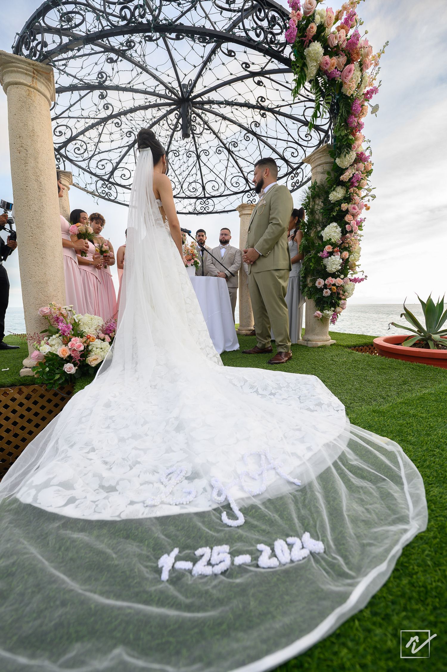Bride in a long lace wedding veil embroidered with the date 7-25-2021 faces the groom and bridesmaids at an oceanfront gazebo ceremony.