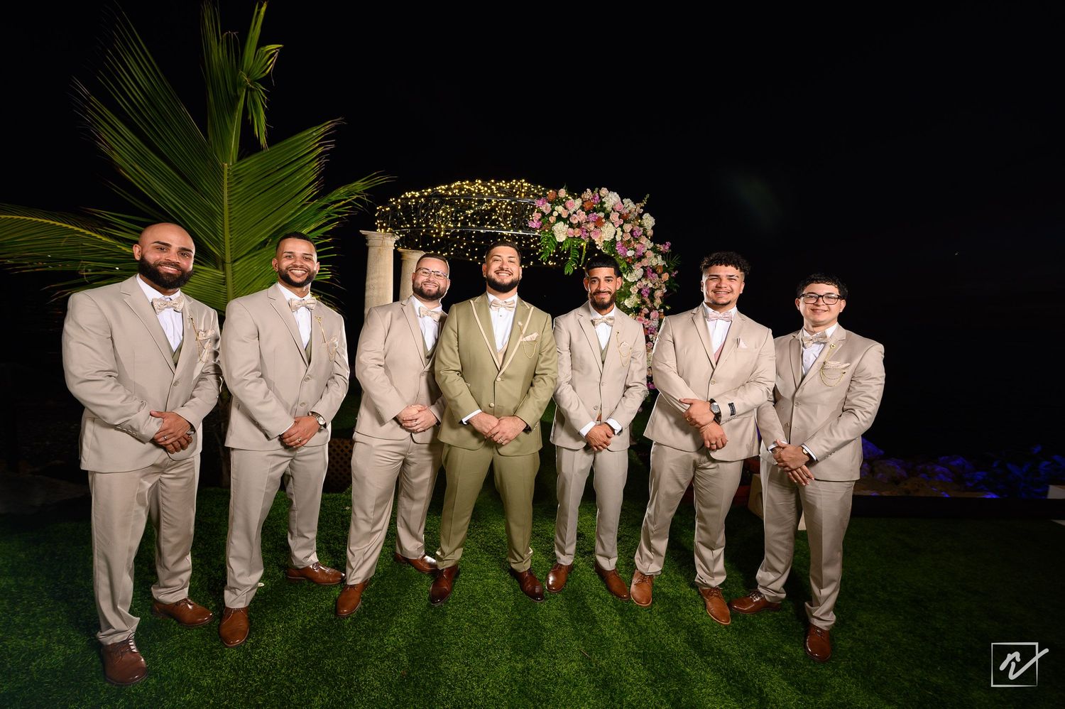 Eight groomsmen in beige suits pose together on a lawn at night in front of a floral wedding arch and palm leaves.
