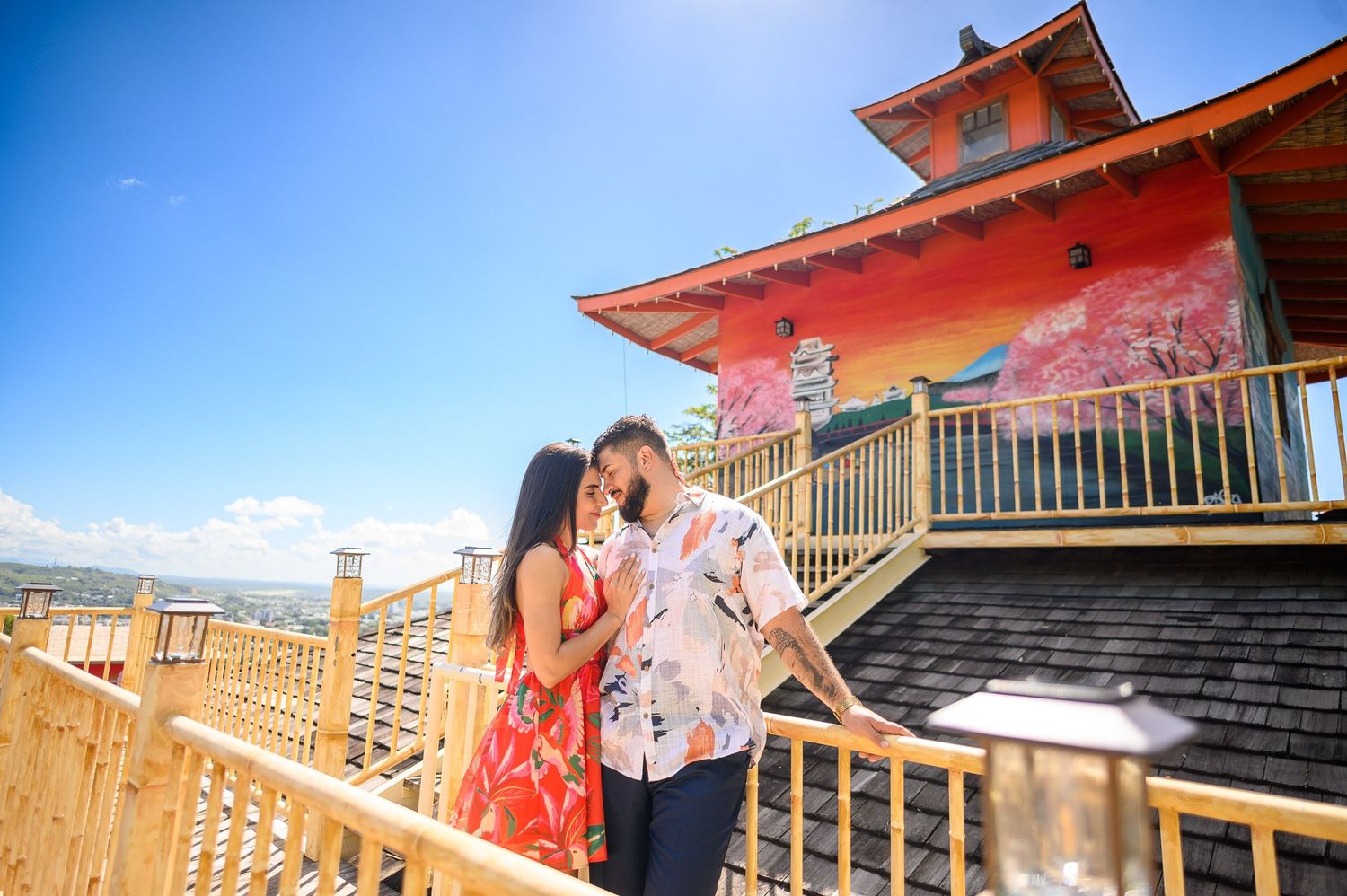 Couple sharing a romantic moment on a wooden deck beside a colorful Asian-style building under a bright blue sky.