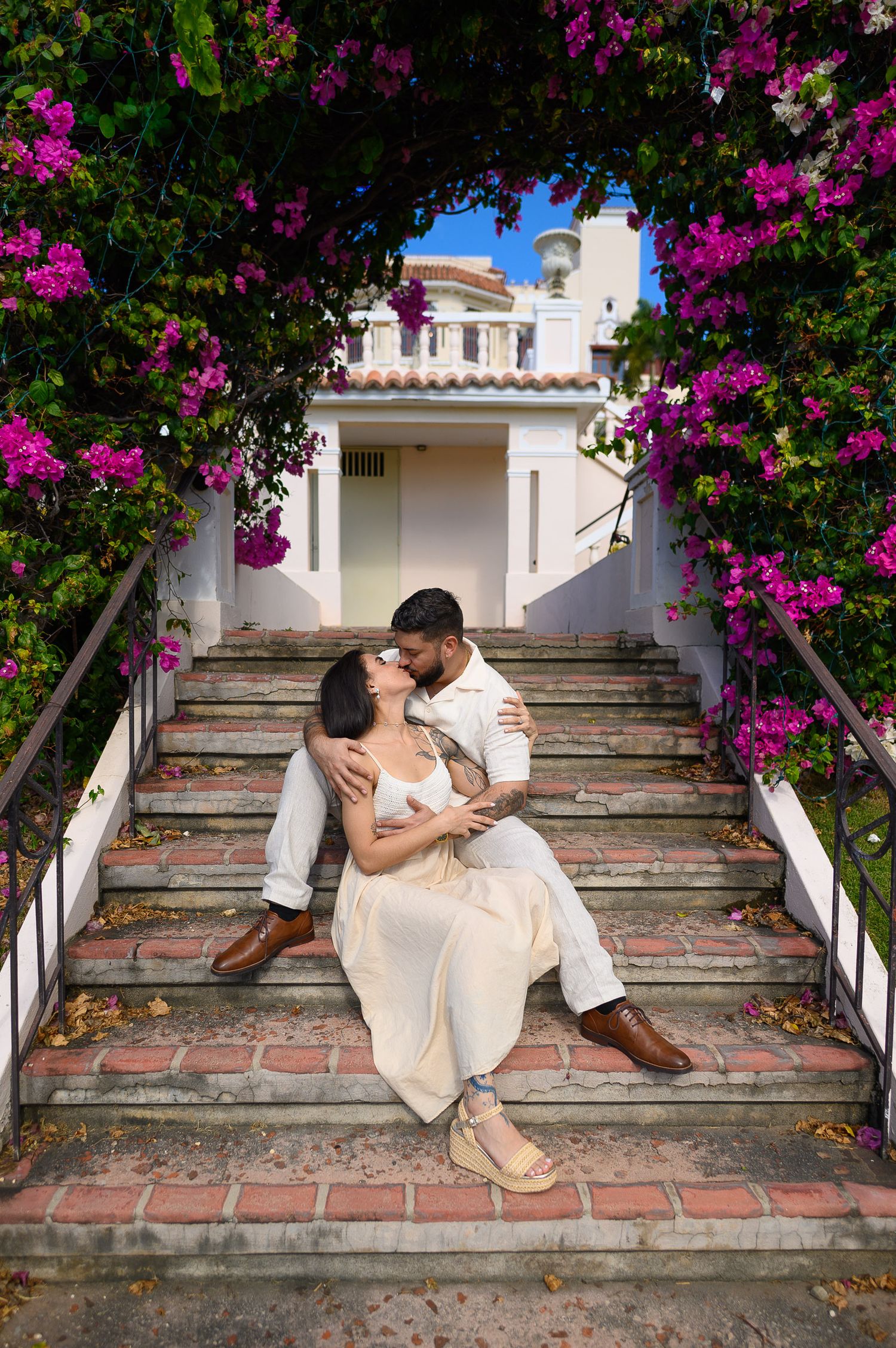 Couple sitting on brick steps under a bougainvillea arch, sharing a romantic moment near a white Mediterranean villa.