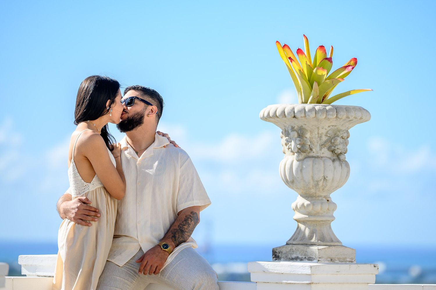 Couple sharing a romantic kiss on a sunny terrace with ocean views and a decorative stone urn with tropical flowers.