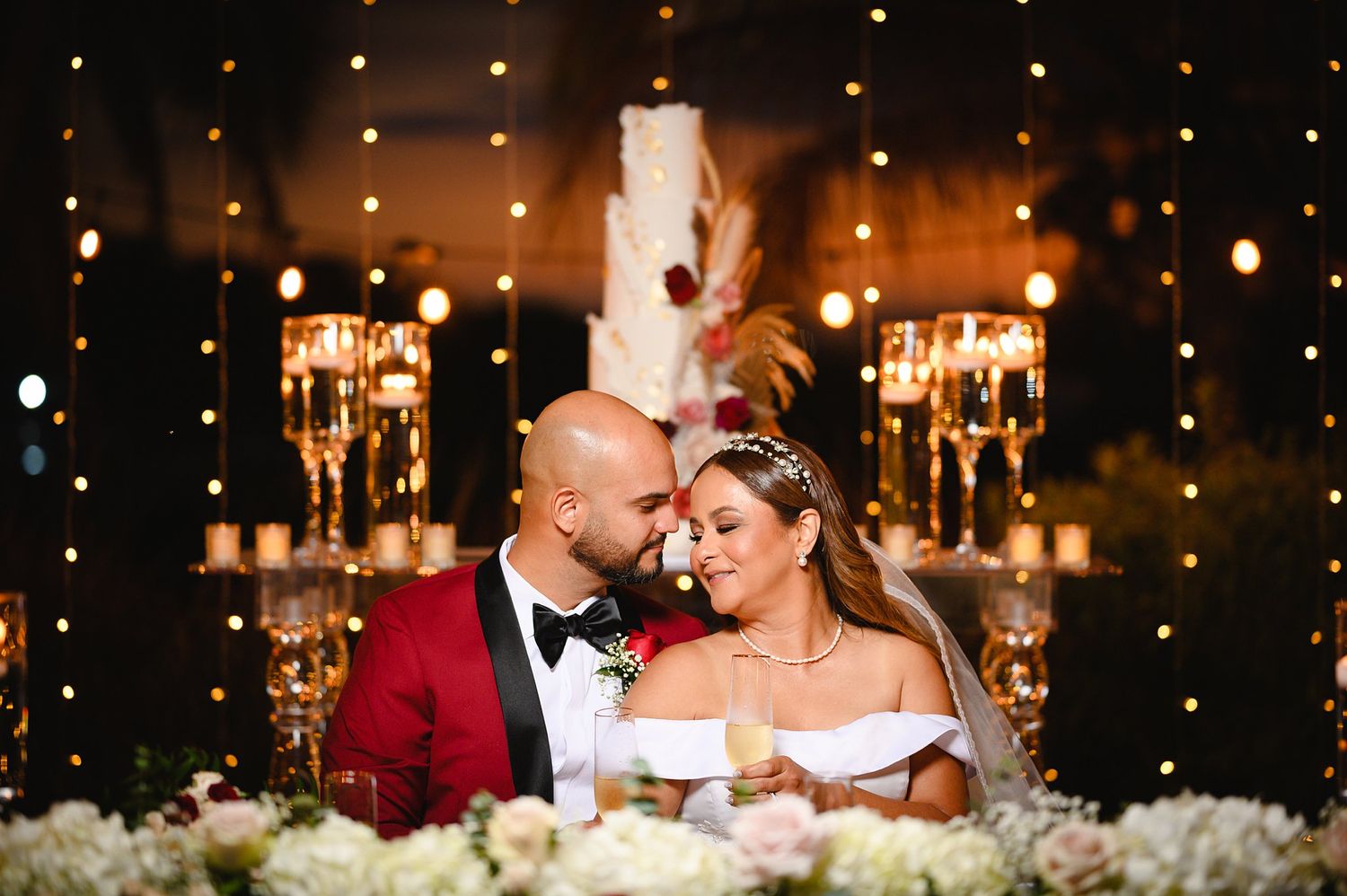 Bride and groom share romantic moment at wedding reception sweetheart table with fairy lights and cake backdrop.