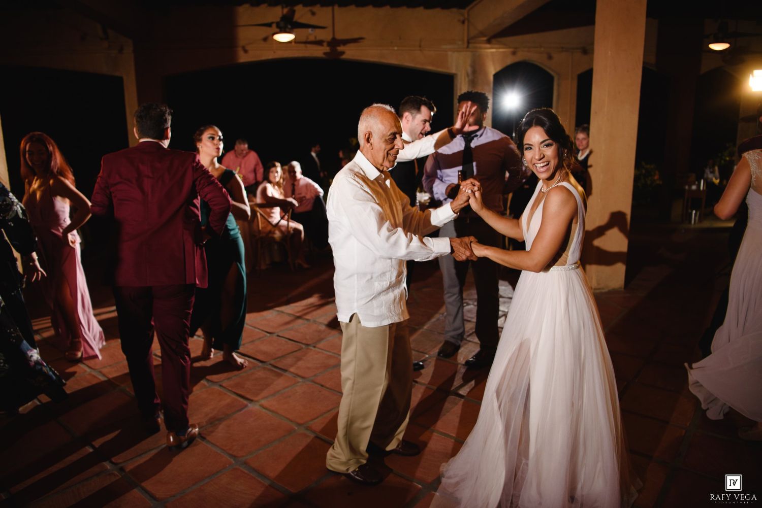 Bride in white gown dances with older man at outdoor wedding reception while guests celebrate on terrace at night.