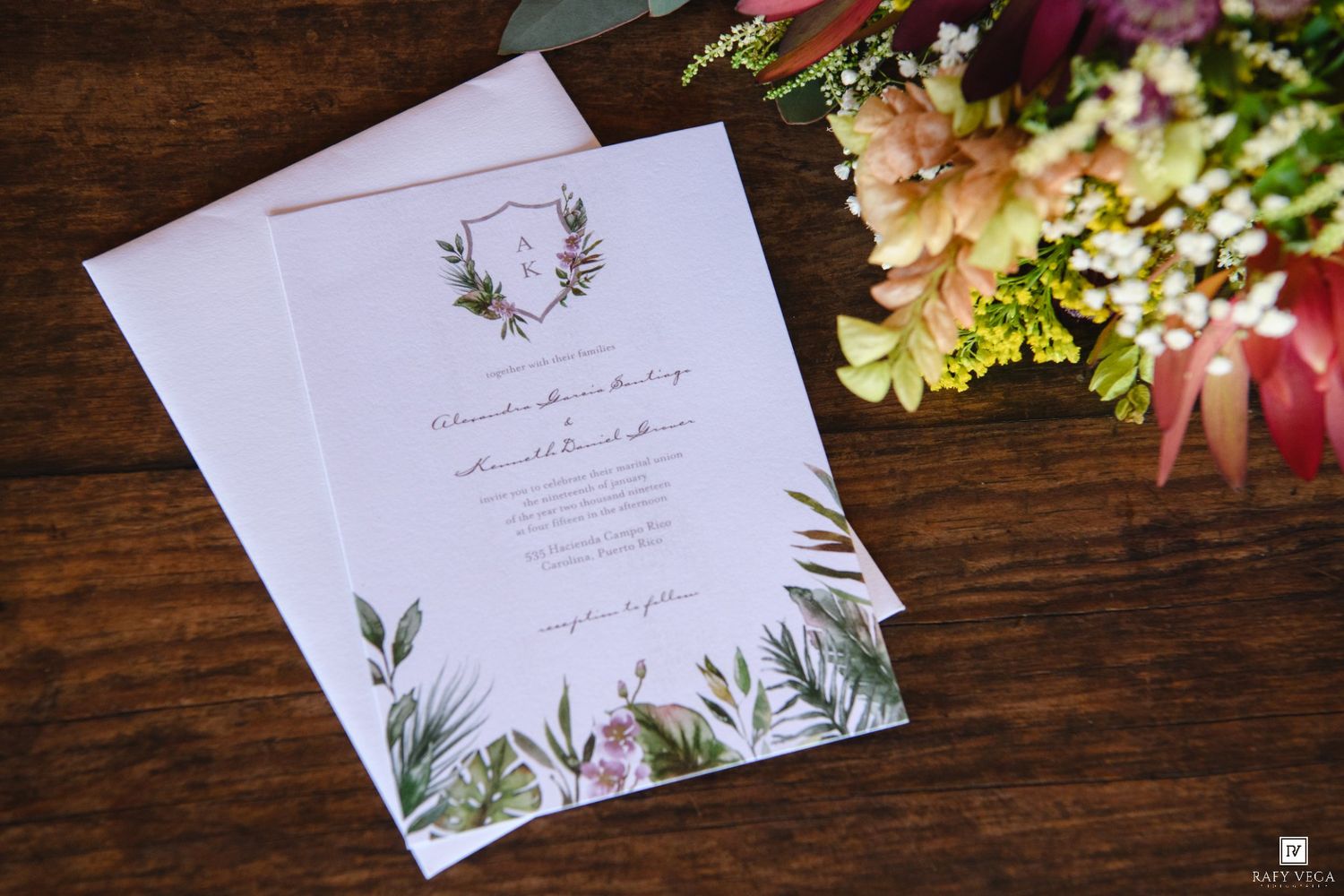 Elegant botanical wedding invitation with greenery border and monogram crest on rustic wood table beside colorful floral bouquet.