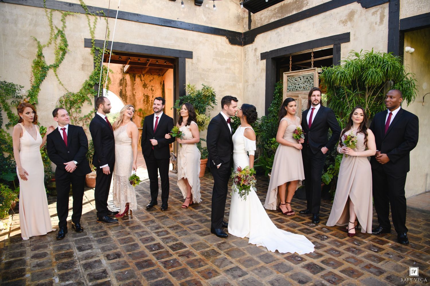 Wedding party poses outdoors in courtyard, bridesmaids in blush gowns and groomsmen in black suits flanking bride and groom.